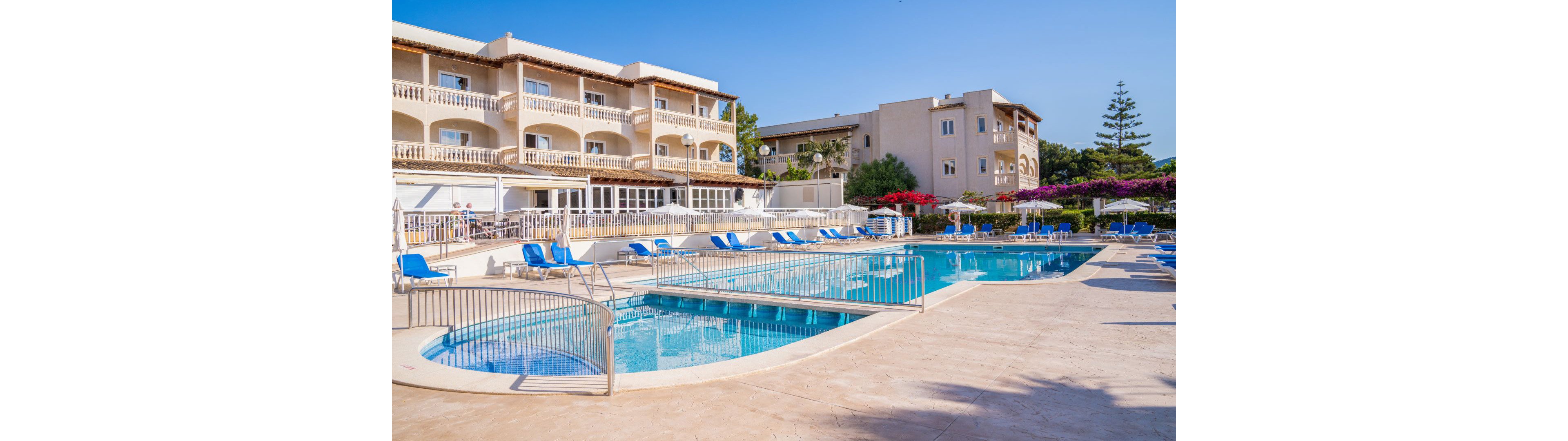 A spacious outdoor swimming pool is featured in front of a multi-story resort hotel. Blue lounge chairs and umbrellas line the poolside, creating a relaxing vacation atmosphere. The setting is bright and sunny, with clear blue skies and Mediterranean-style architecture. No visible people or text are present in the image.