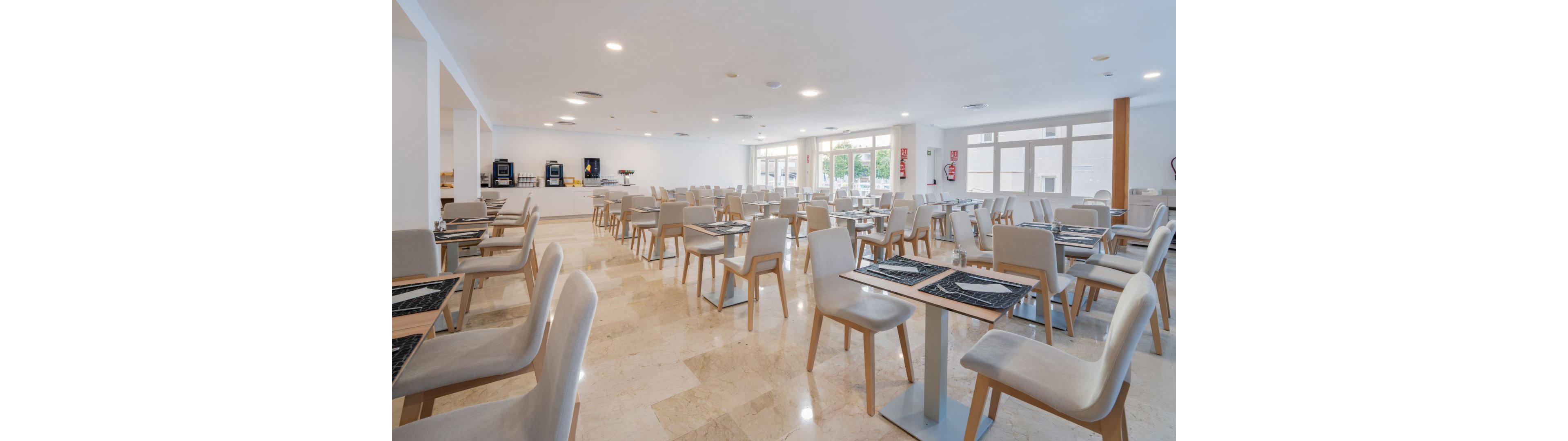 A spacious, contemporary dining room features neatly arranged tables and chairs, all unoccupied. The setting is bright with large windows allowing ample daylight to fill the space. Neutral tones dominate the palette, creating a clean and inviting atmosphere. Beverage machines are visible in the background, enhancing the modern cafeteria or restaurant feel.