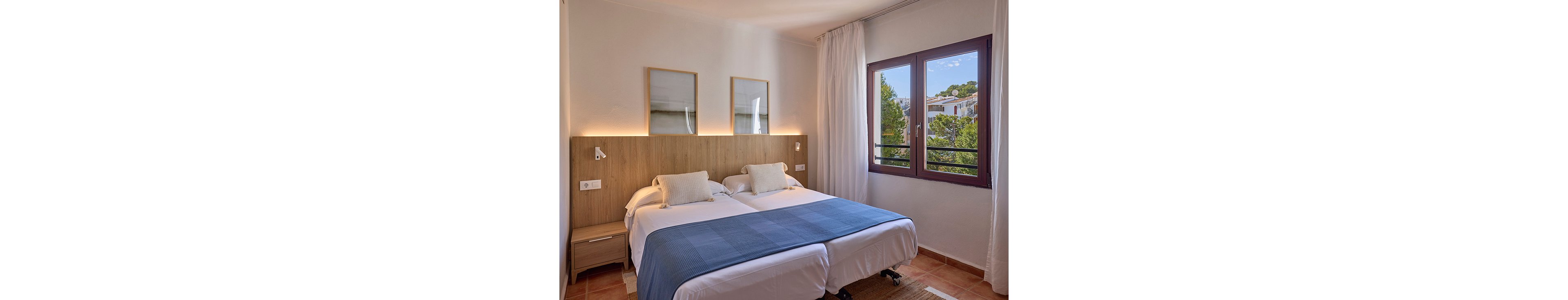 A modern hotel bedroom features two neatly made twin beds pushed together with a shared wooden headboard. Soft white bedding is accented by a blue runner and matching pillows, creating a calm and tidy atmosphere. Warm wall lighting, simple framed artwork, and bedside tables with reading lamps enhance the cozy interior. A large window with sheer curtains reveals trees and nearby buildings under a clear sky, filling the room with natural light.