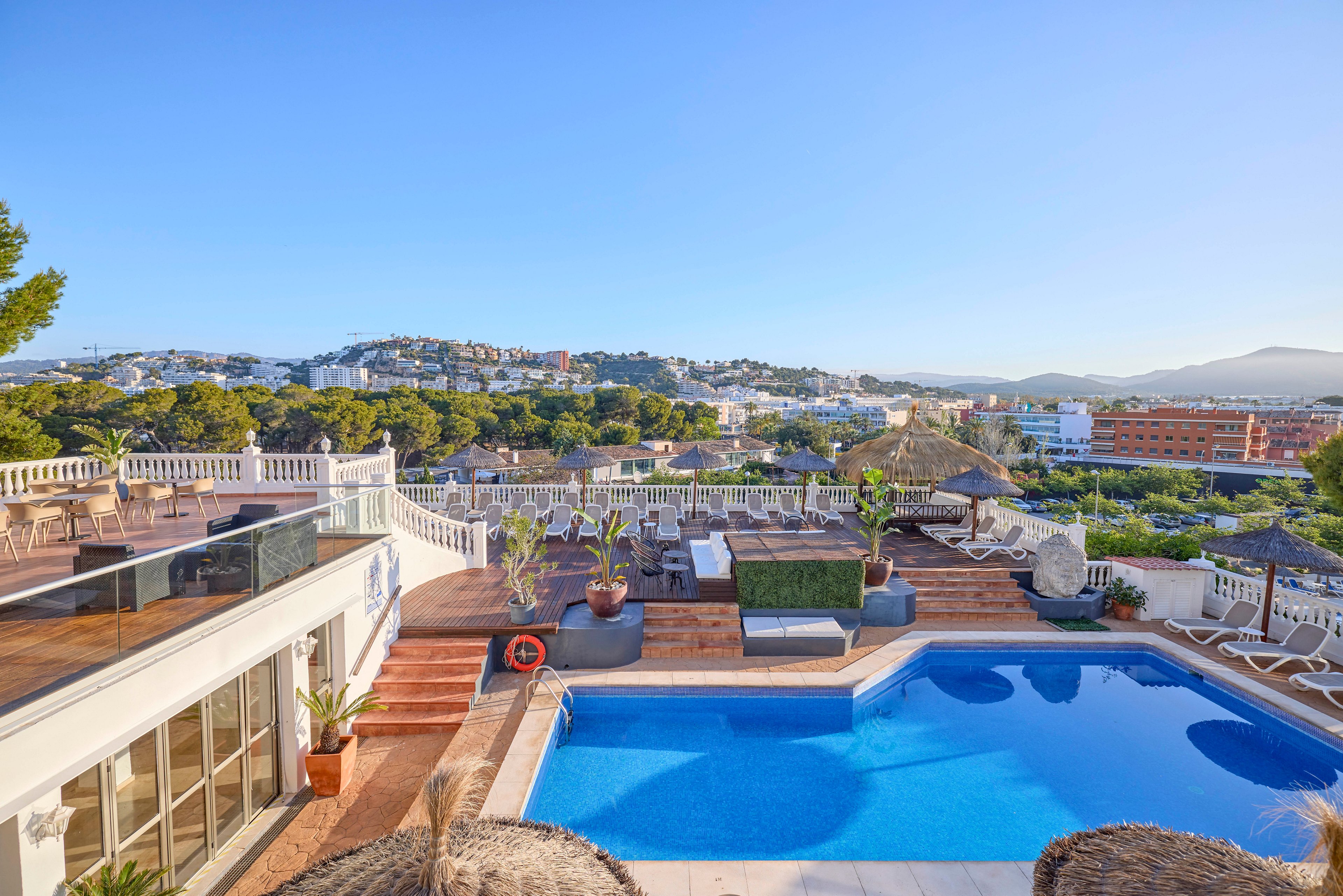A bright blue rooftop swimming pool sits atop a terraced building with lounge chairs and seating areas. The scene overlooks a coastal town with white buildings, green hills, and distant mountains under a clear blue sky. Sunlit railings, potted plants, and thatched umbrellas add a relaxed resort atmosphere. The calm water and wide view suggest a peaceful vacation setting.