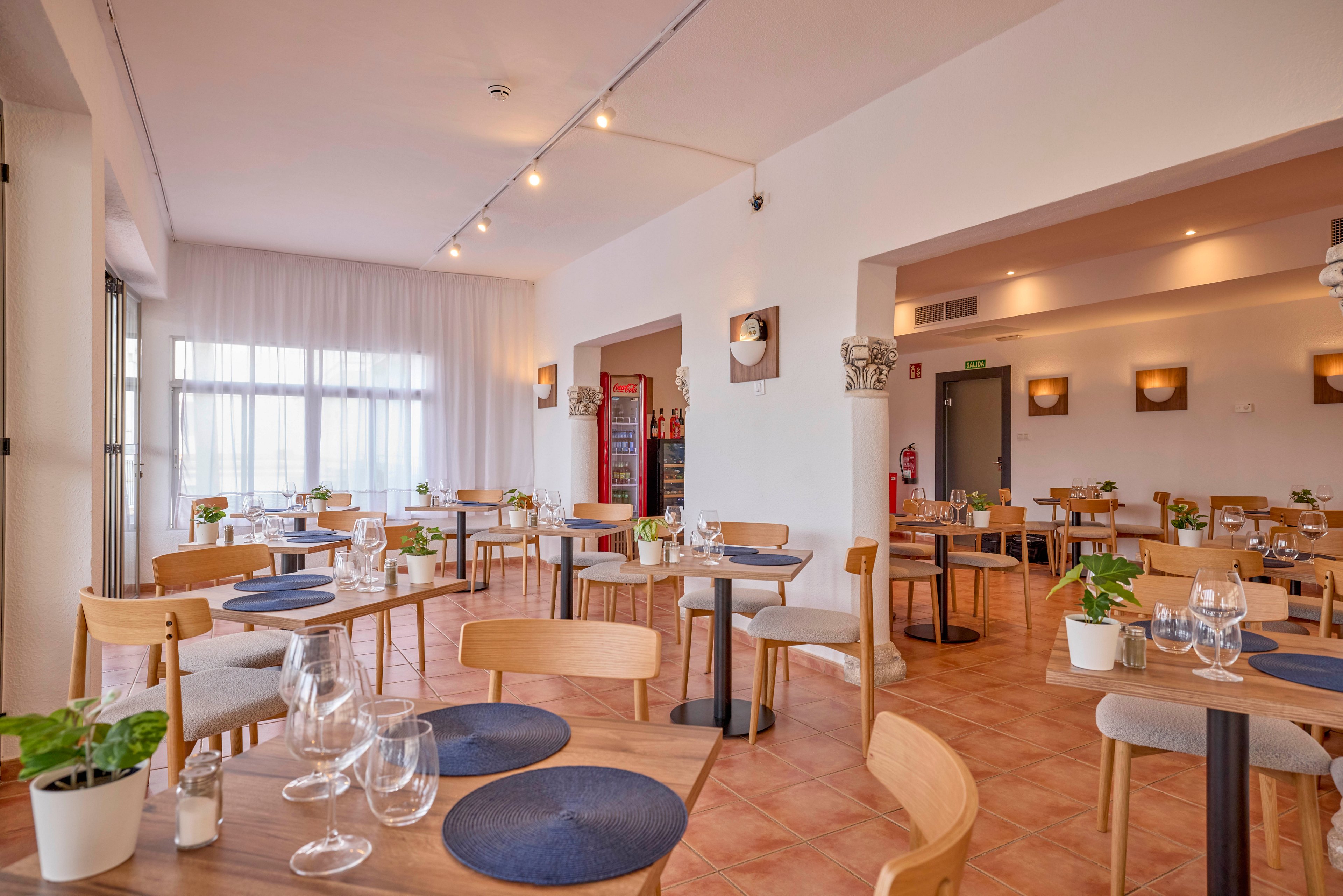 A bright, modern restaurant dining room is set with wooden tables and chairs. Each table features blue round placemats, clear wine glasses, and small potted plants as centerpieces. Large windows with sheer white curtains allow natural light to fill the space, highlighting the warm tiled floor. Wall sconces and ceiling track lights create a cozy yet contemporary atmosphere in the empty dining area.