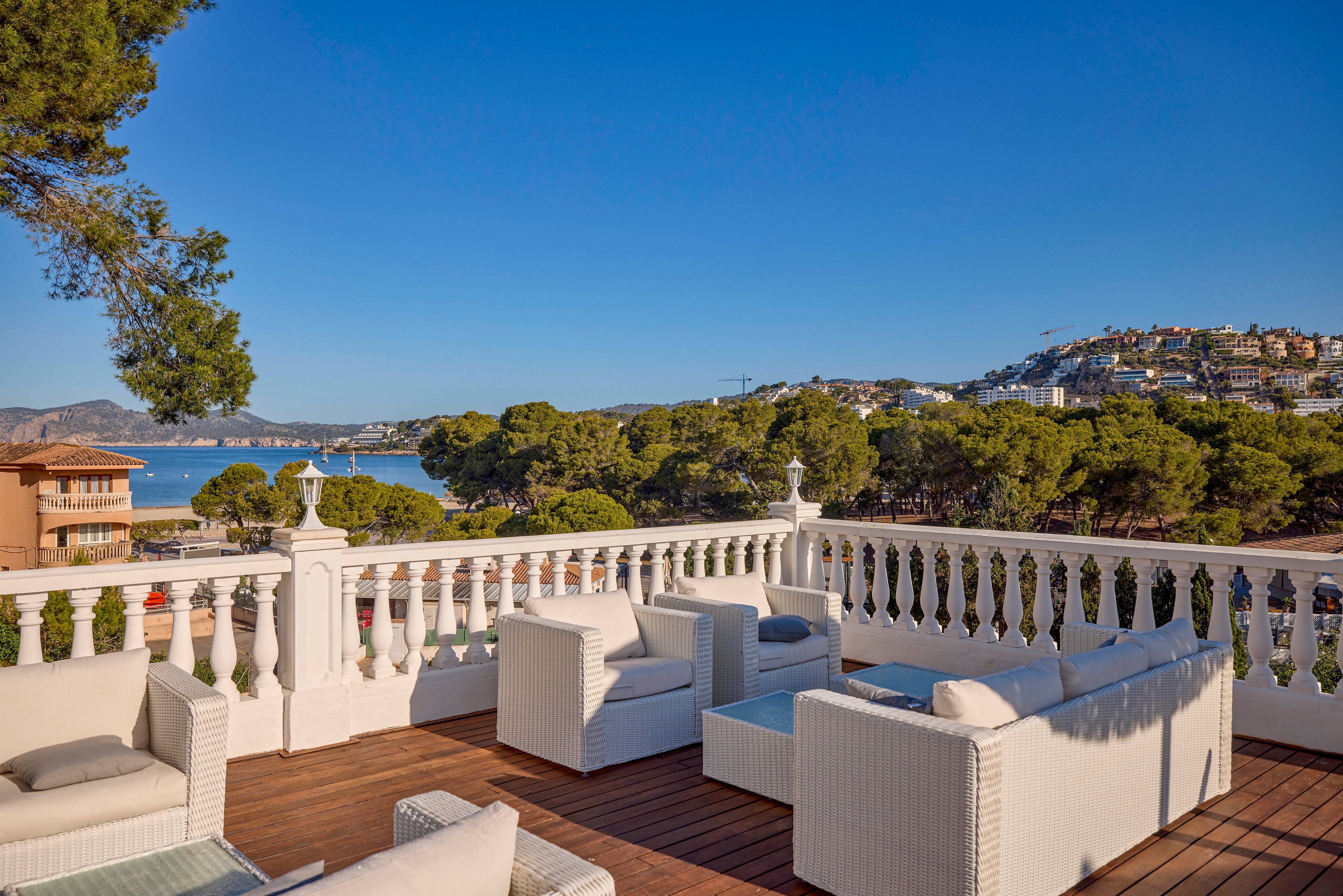 A spacious outdoor terrace with white wicker lounge furniture overlooks a scenic coastal bay. The wooden deck is bordered by a classic white balustrade, framing views of green trees and blue water. In the distance, hillside buildings and villas rise above the shoreline under a clear blue sky. The scene suggests a relaxed, luxurious setting ideal for leisure and socializing.
