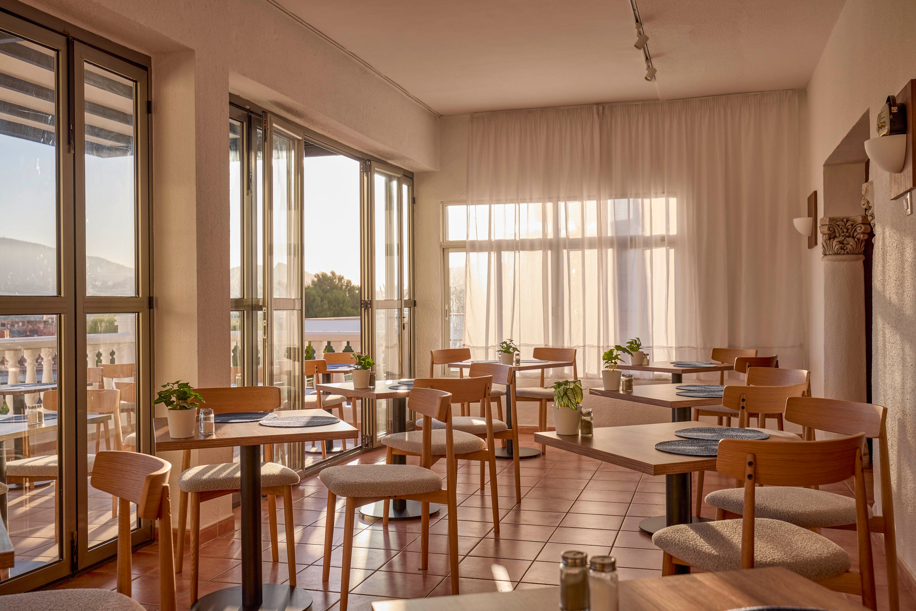 A bright, modern cafe interior is filled with natural light streaming through large floor-to-ceiling windows. Simple wooden tables and chairs are neatly arranged on a tiled floor, with small plants placed as centerpieces. Sheer curtains soften the sunlight and create a warm, inviting atmosphere. Outside, a terrace with additional seating overlooks a scenic view, enhancing the relaxed dining experience.