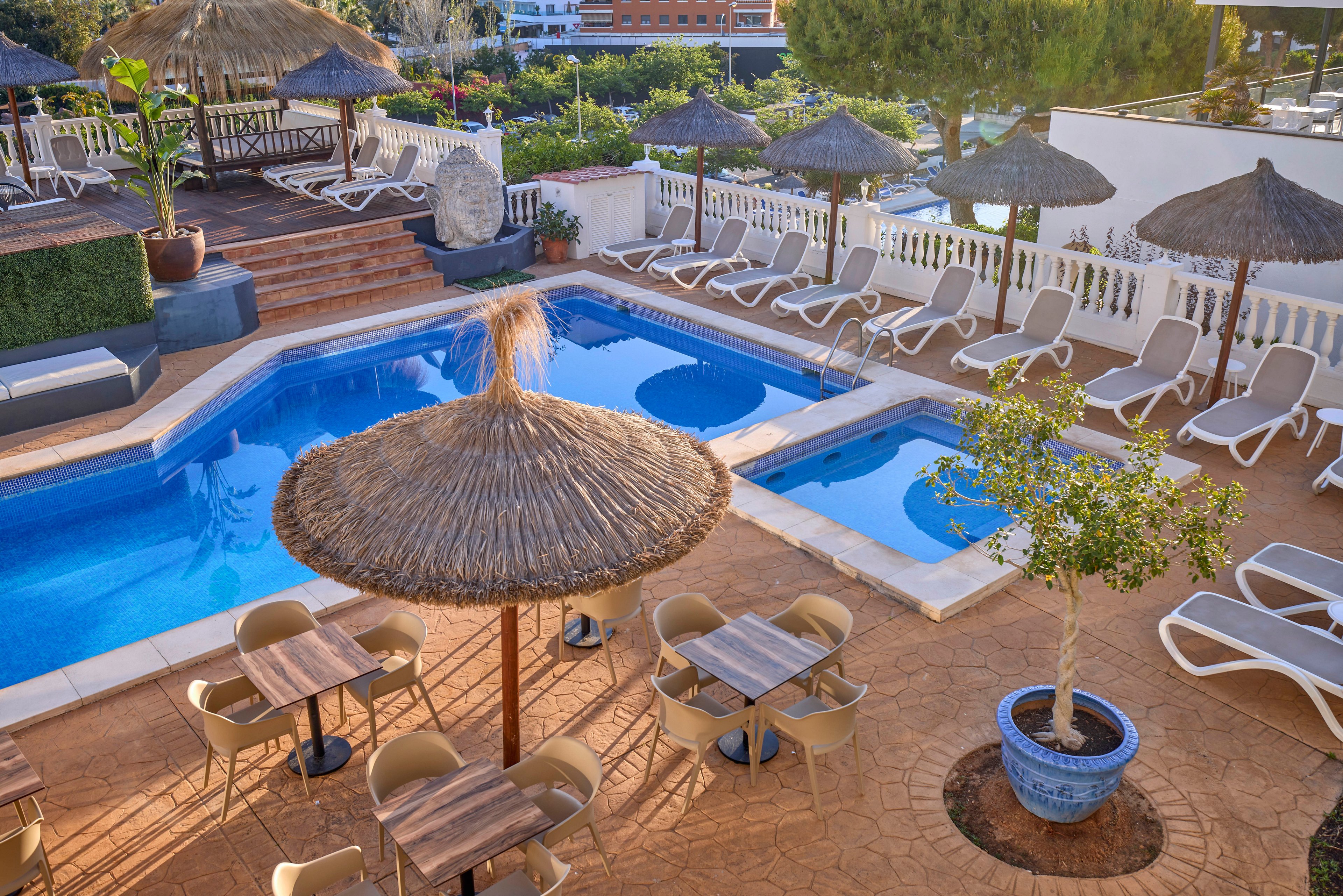 An outdoor resort pool area features a main swimming pool and a smaller adjacent pool surrounded by white sun loungers. Straw parasols shade several tables and chairs on a terracotta-tiled terrace. A raised wooden deck with additional seating and thatched umbrellas overlooks the water. Lush greenery, potted plants, and distant buildings frame the relaxing vacation setting. The scene is brightly lit, suggesting a warm, sunny day ideal for leisure.
