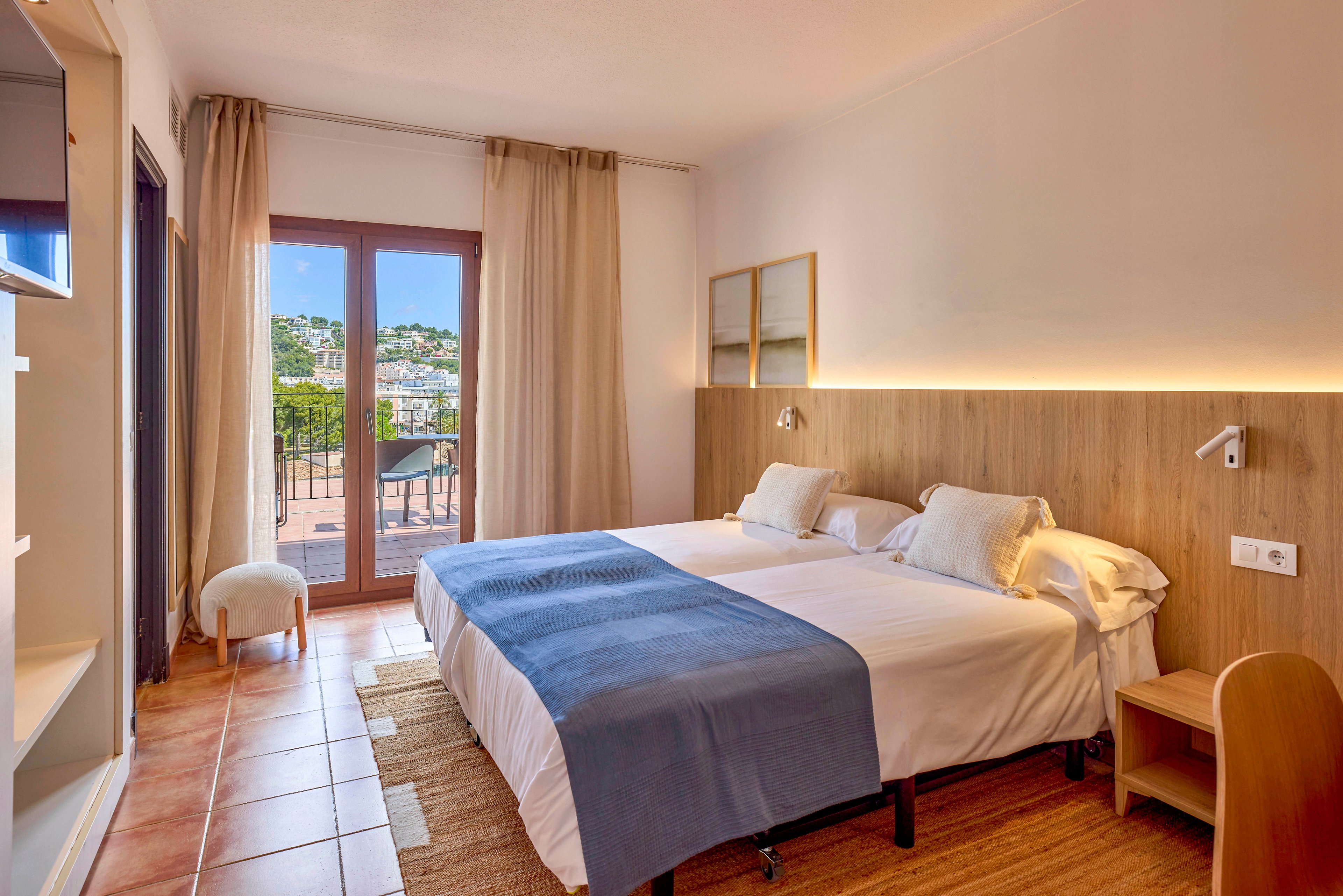 A cozy hotel room features two neatly made twin beds with white linens and blue throws against a wooden headboard. Warm lighting and neutral curtains create a calm, inviting atmosphere. A glass door opens to a balcony with a view of a hillside town in the distance. Simple furniture and soft tones emphasize a minimalist, modern design ideal for relaxation.