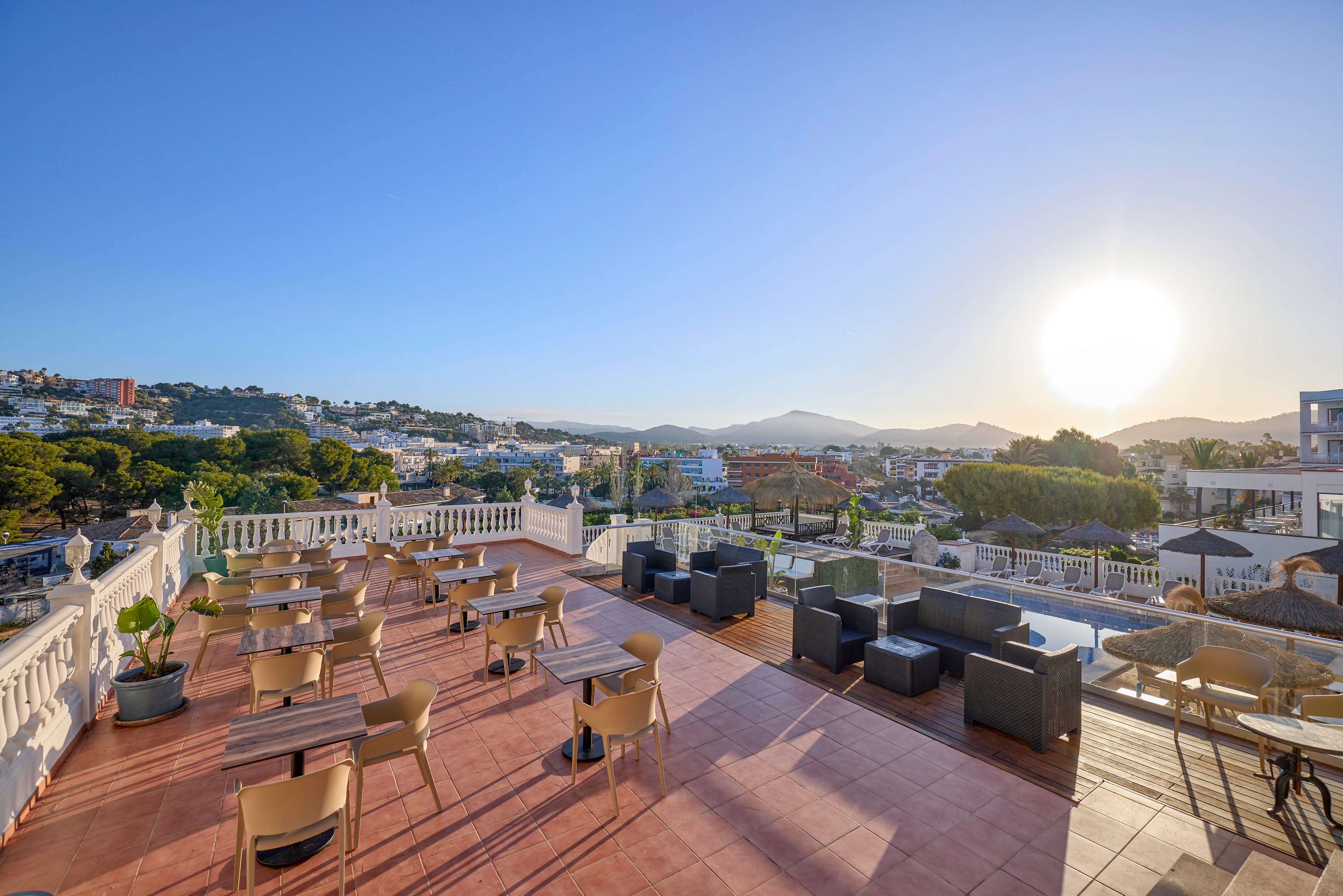 A spacious rooftop terrace with tables and chairs overlooks a hillside town under a clear blue sky. The sun is low on the horizon, casting warm light across the tiled floor and outdoor seating area. Modern lounge furniture is arranged along the edge, offering views of distant mountains and surrounding buildings. The scene suggests a relaxed, open-air setting ideal for dining or social gatherings.