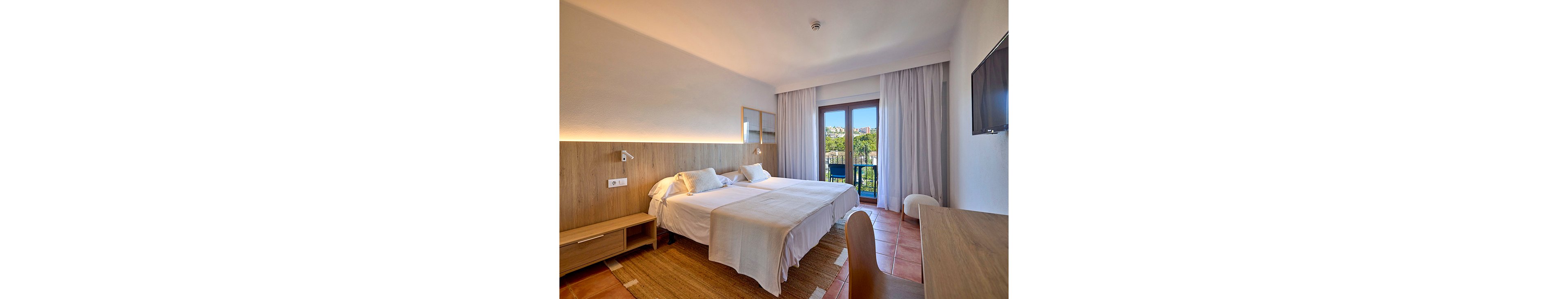 A contemporary hotel room features two neatly made twin beds with white linens and a wooden headboard. Warm ambient lighting runs along the wall, creating a cozy and inviting atmosphere. A flat-screen television is mounted on the wall opposite a simple desk and chair. Large glass doors with sheer curtains open to a balcony, revealing greenery and buildings outside.