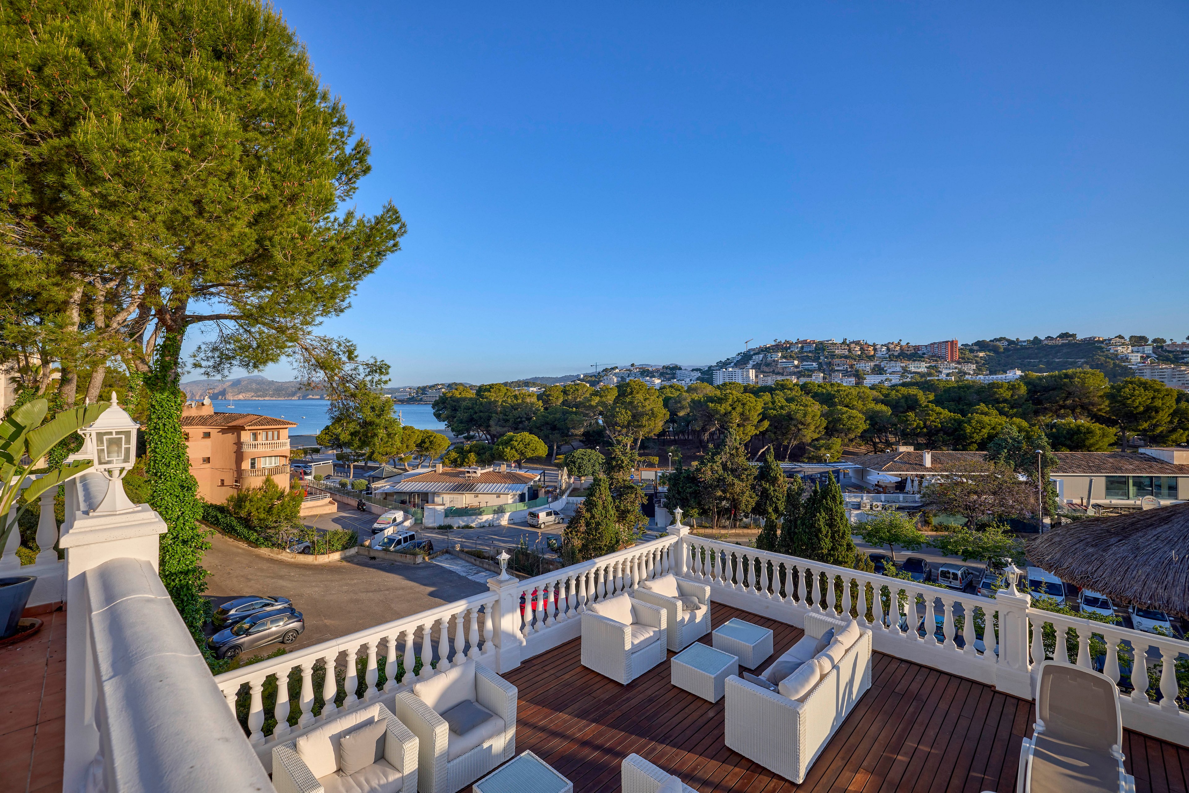 An elevated terrace lounge with modern white outdoor furniture overlooks a coastal town. The scene includes trees, residential buildings, and a glimpse of the sea in the distance. Warm sunlight and clear blue skies create a relaxed, vacation-like atmosphere. Decorative railings and potted plants add elegance to the open-air seating area.