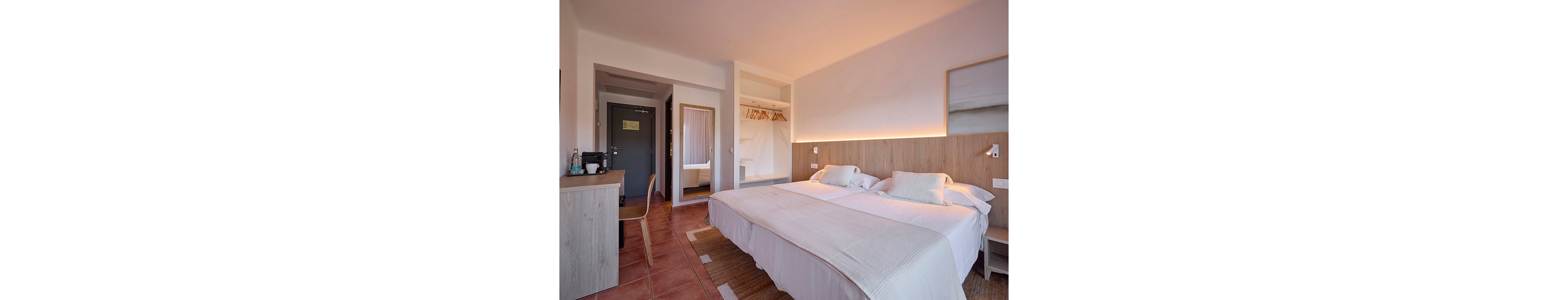 A bright, modern hotel room features two neatly made twin beds pushed together with white linens and soft pillows. The space includes a wooden desk with a chair, a mirror, and an open wardrobe with hangers. Warm under-bed and headboard lighting create a cozy atmosphere against neutral walls and tiled flooring. A doorway leads to the bathroom, adding depth and context to the interior scene.