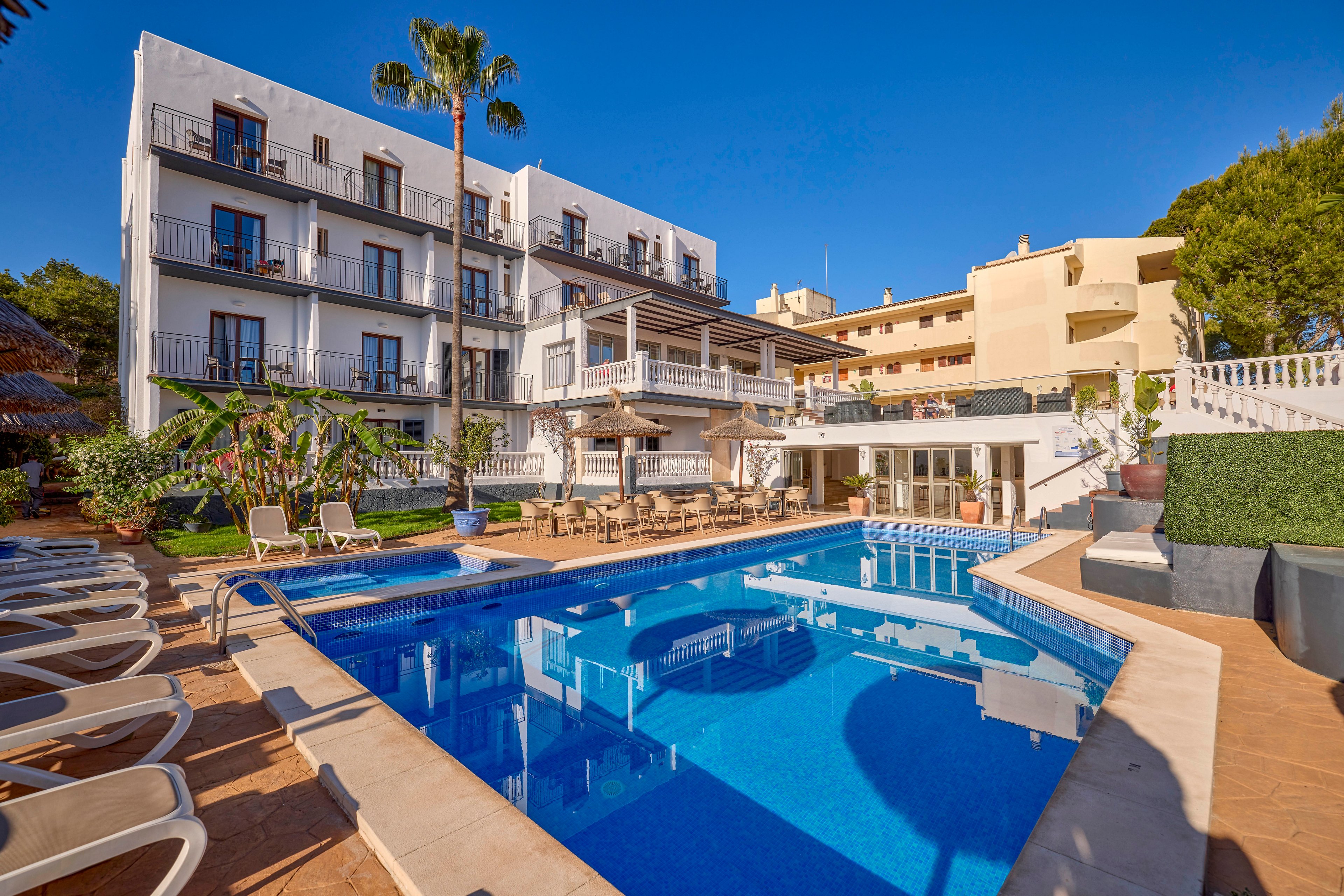 A bright hotel courtyard features a clear blue outdoor swimming pool surrounded by sun loungers. The multi-story white building has balconies overlooking the pool area and a shaded terrace with tables and chairs. Palm trees and lush plants add a tropical touch to the scene. Warm sunlight and clear skies create a relaxed vacation atmosphere.