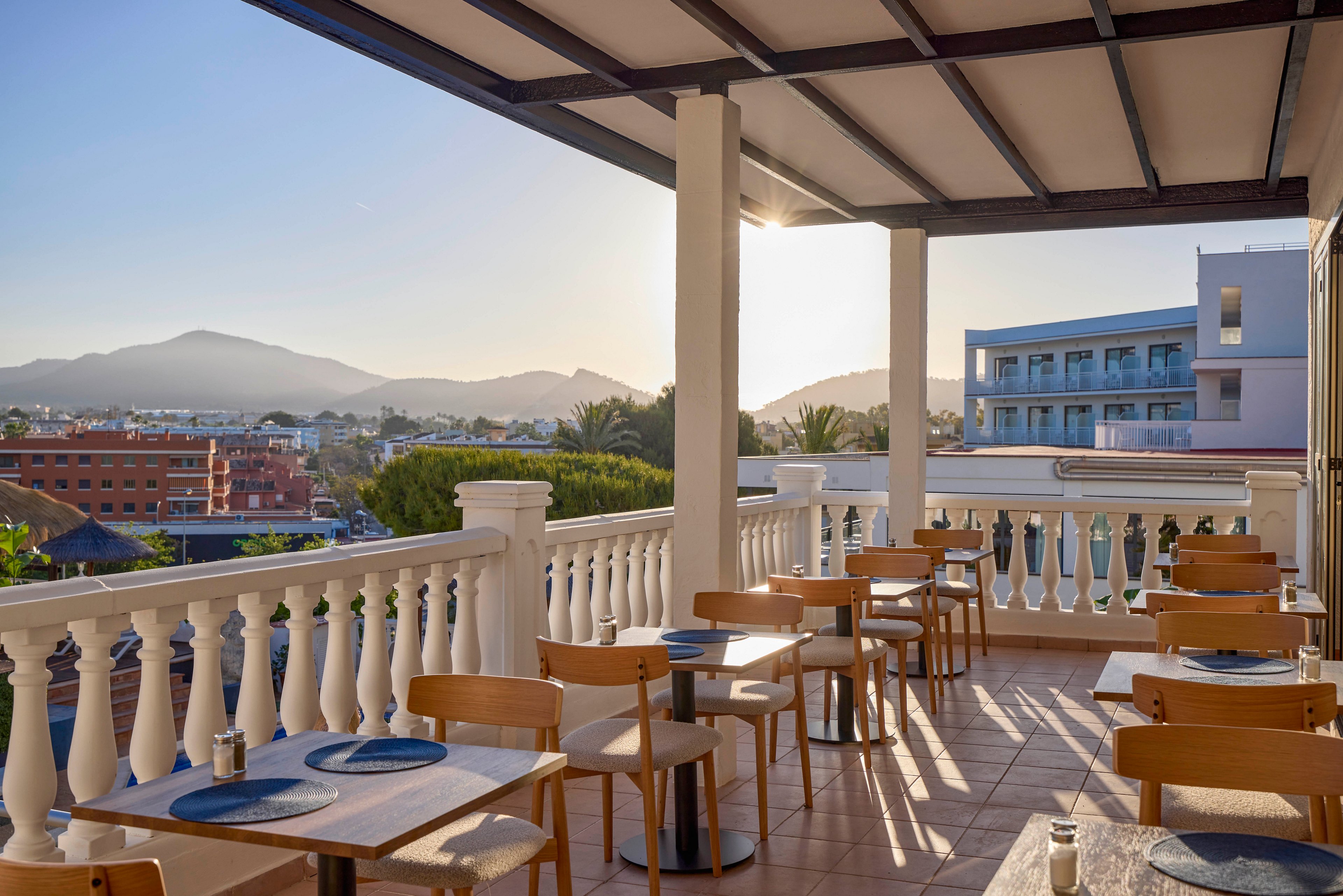 An outdoor restaurant terrace is set with wooden tables and chairs under a covered pergola. Blue placemats and simple table settings create a clean, modern dining atmosphere. The terrace overlooks nearby buildings, trees, and distant mountains in the warm light of either sunrise or sunset. Long shadows and clear skies contribute to a calm, inviting ambiance.