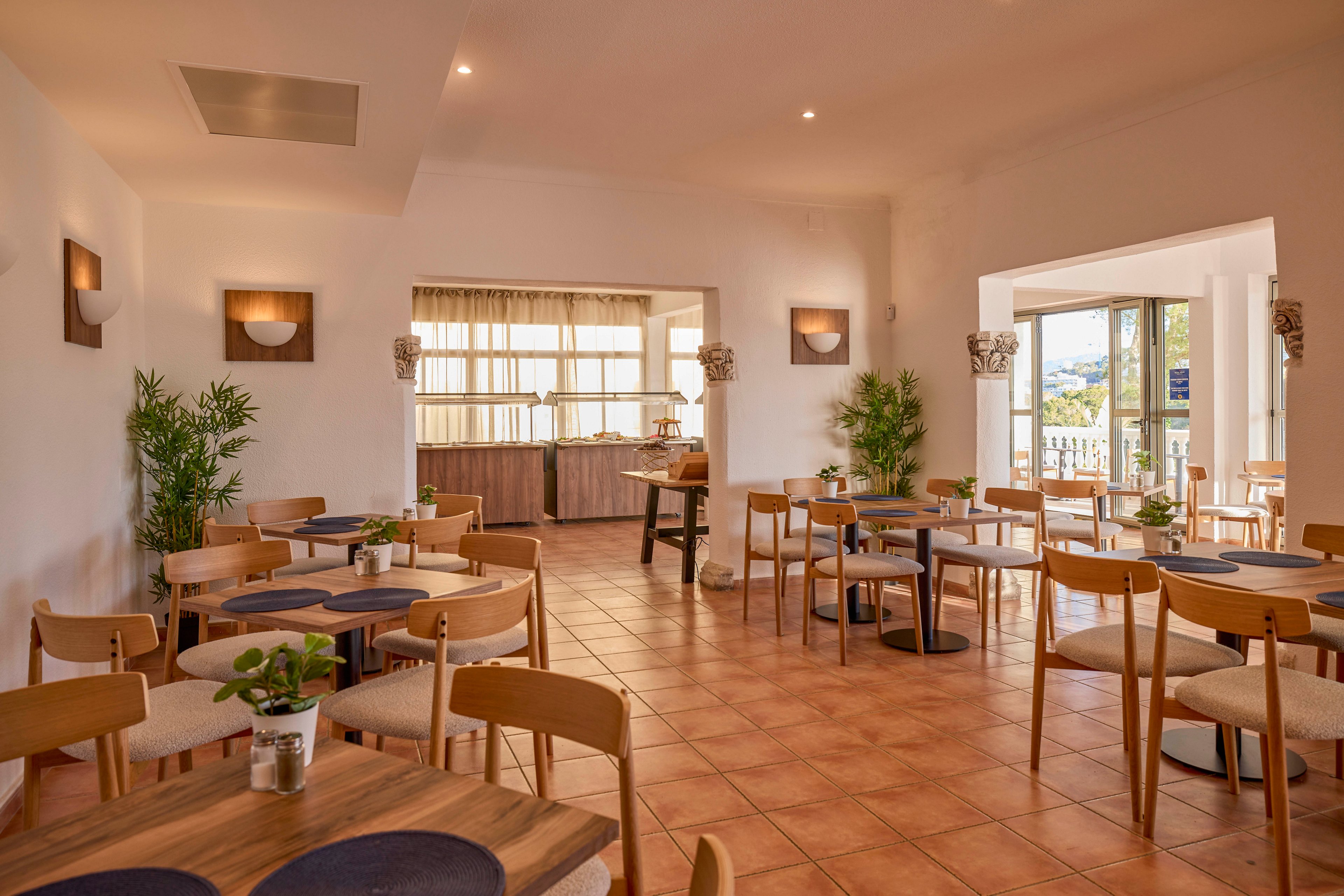 A cozy restaurant dining room features wooden tables and chairs arranged neatly across a tiled floor. Large windows and glass doors let in abundant natural light, revealing a balcony and scenic view outside. Potted plants and simple wall sconces add warmth and a minimalist decorative touch. The overall atmosphere is calm, inviting, and well-suited for casual dining or small gatherings.