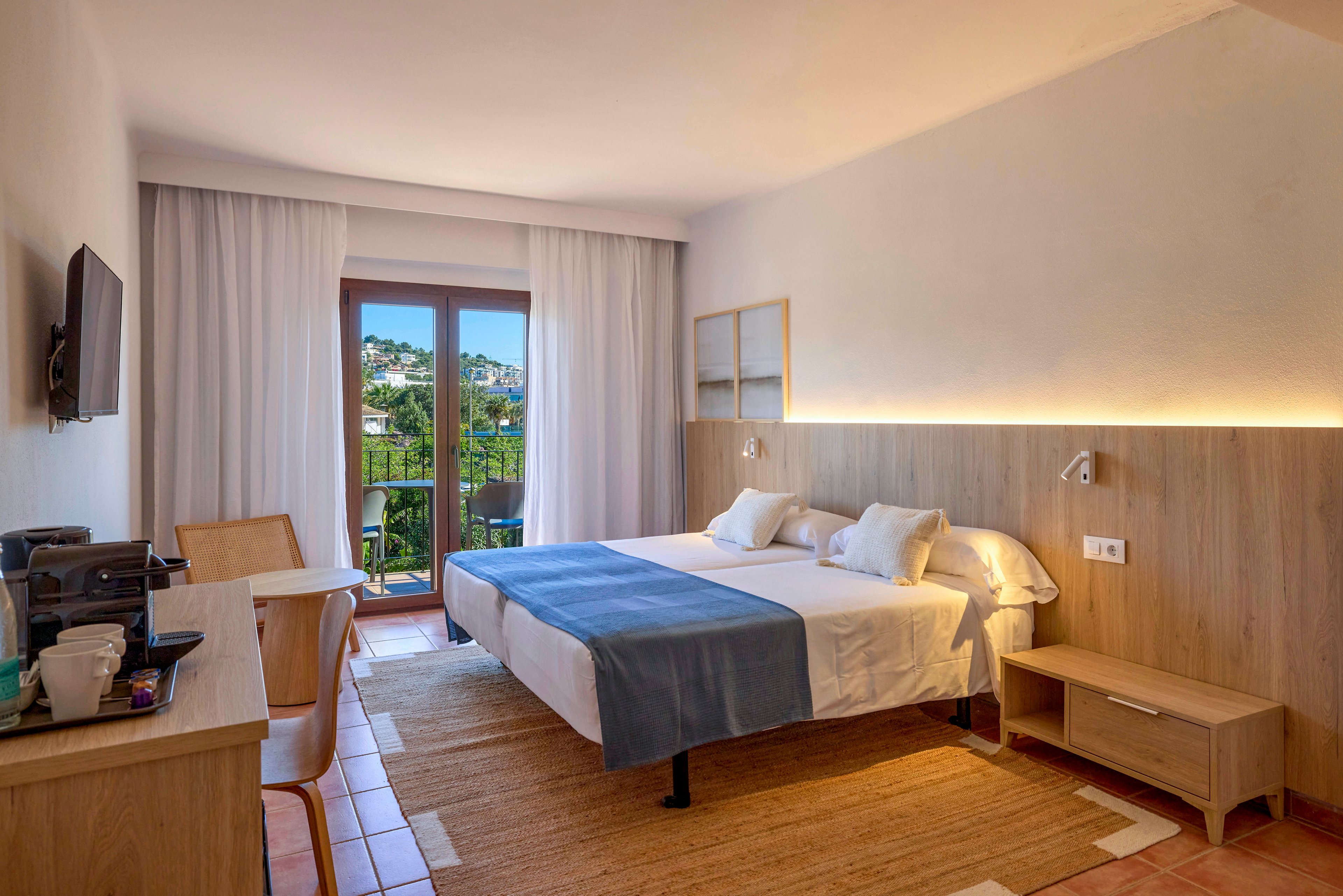 A contemporary hotel room features two single beds pushed together with white linens and blue runners. The space includes a wooden desk with a chair, a wall-mounted television, and bedside tables with reading lights. Large glass doors open onto a balcony overlooking greenery and distant buildings. Warm ambient lighting and neutral tones create a calm, inviting atmosphere.