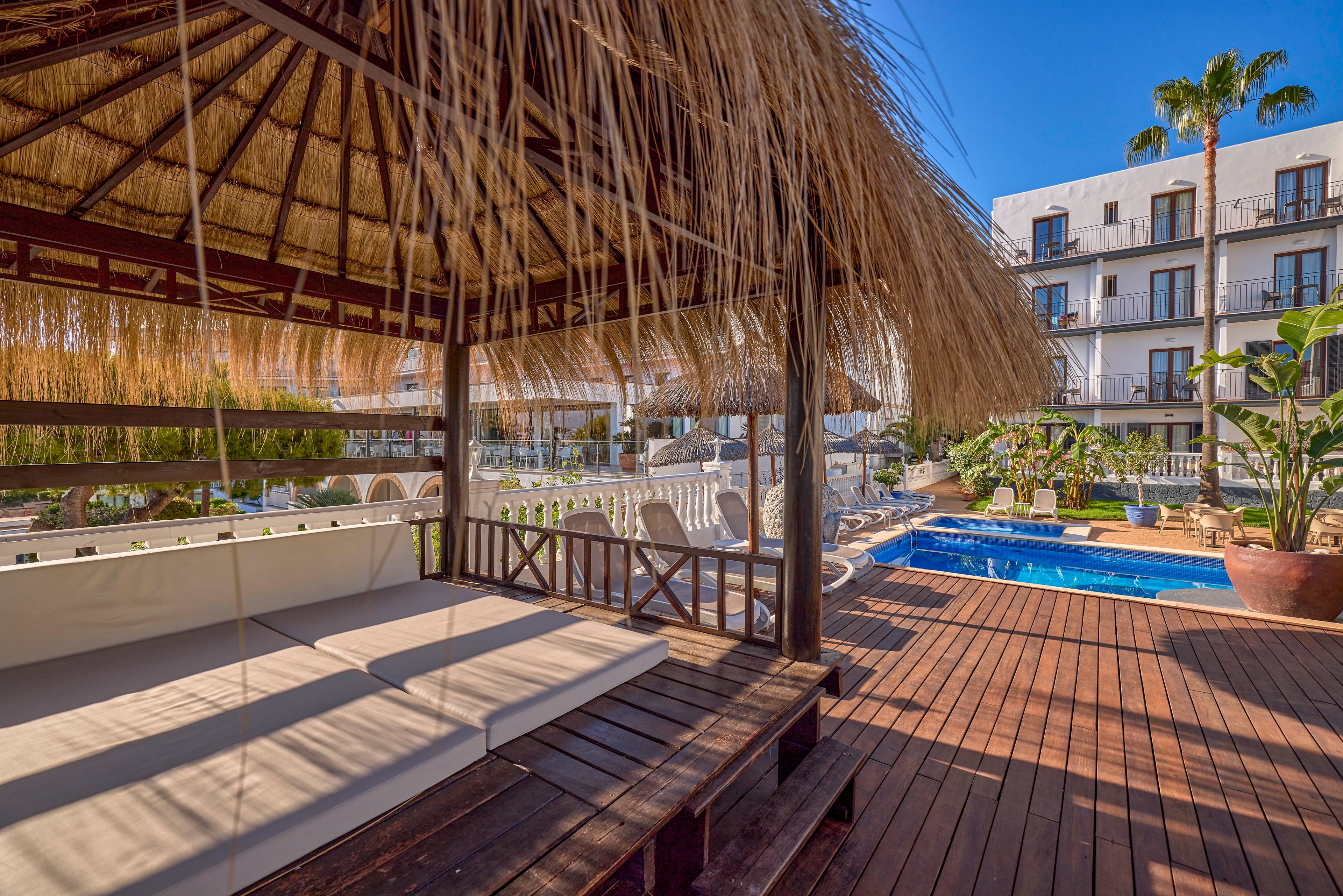 A shaded cabana with cushioned seating sits on a wooden deck beside a small outdoor swimming pool. White lounge chairs line the pool area, surrounded by a white fence and lush tropical plants. A modern hotel building with balconies rises in the background under a clear blue sky. The scene suggests a relaxing vacation atmosphere in a warm, sunny climate.