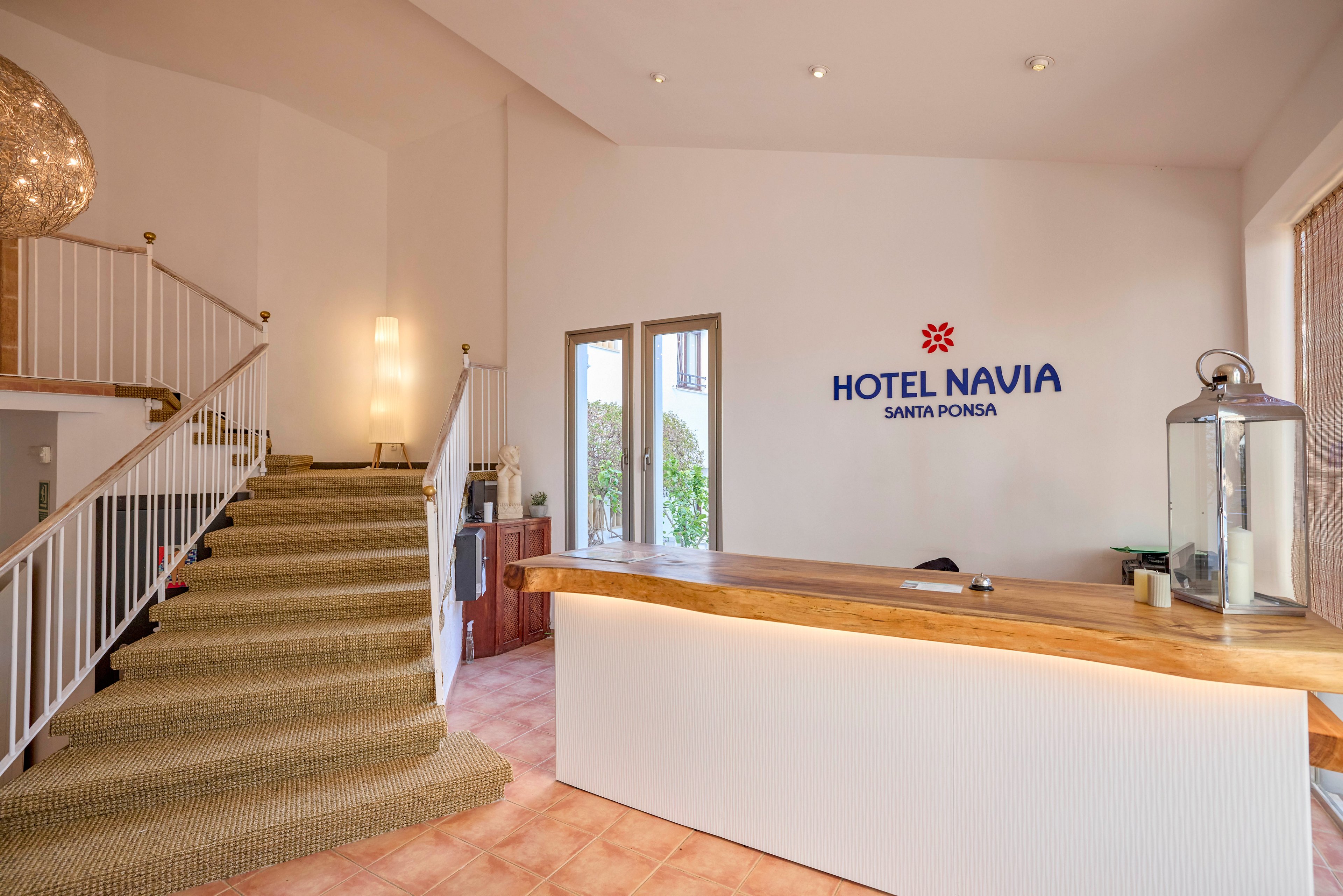 A modern hotel reception area features a long wooden front desk with a white base. To the left, a carpeted staircase with a white railing leads to an upper level. Natural light enters through glass doors at the back, illuminating the warm tiled floor and neutral walls. A large lantern and small decorative items sit on the counter, adding a welcoming touch. The wall behind the desk displays the text "HOTEL NAVIA SANTA PONSA" with a red floral symbol.