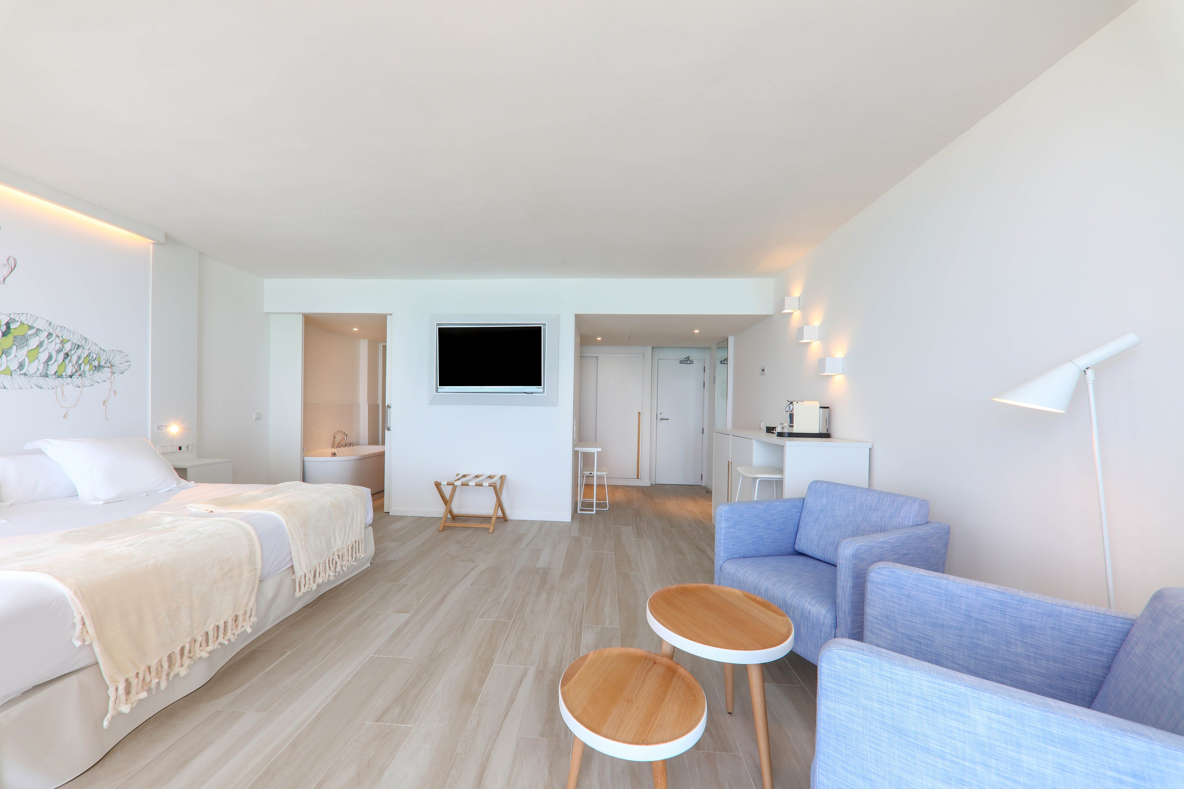 A spacious hotel room features a large bed with neutral bedding, a wall-mounted television, and a cozy seating area with two blue armchairs and round wooden tables. The setting is bright and airy, with light wood flooring and white walls, creating a calm and inviting atmosphere. The room includes a small kitchenette and soft accent lighting, enhancing the modern minimalist style.
