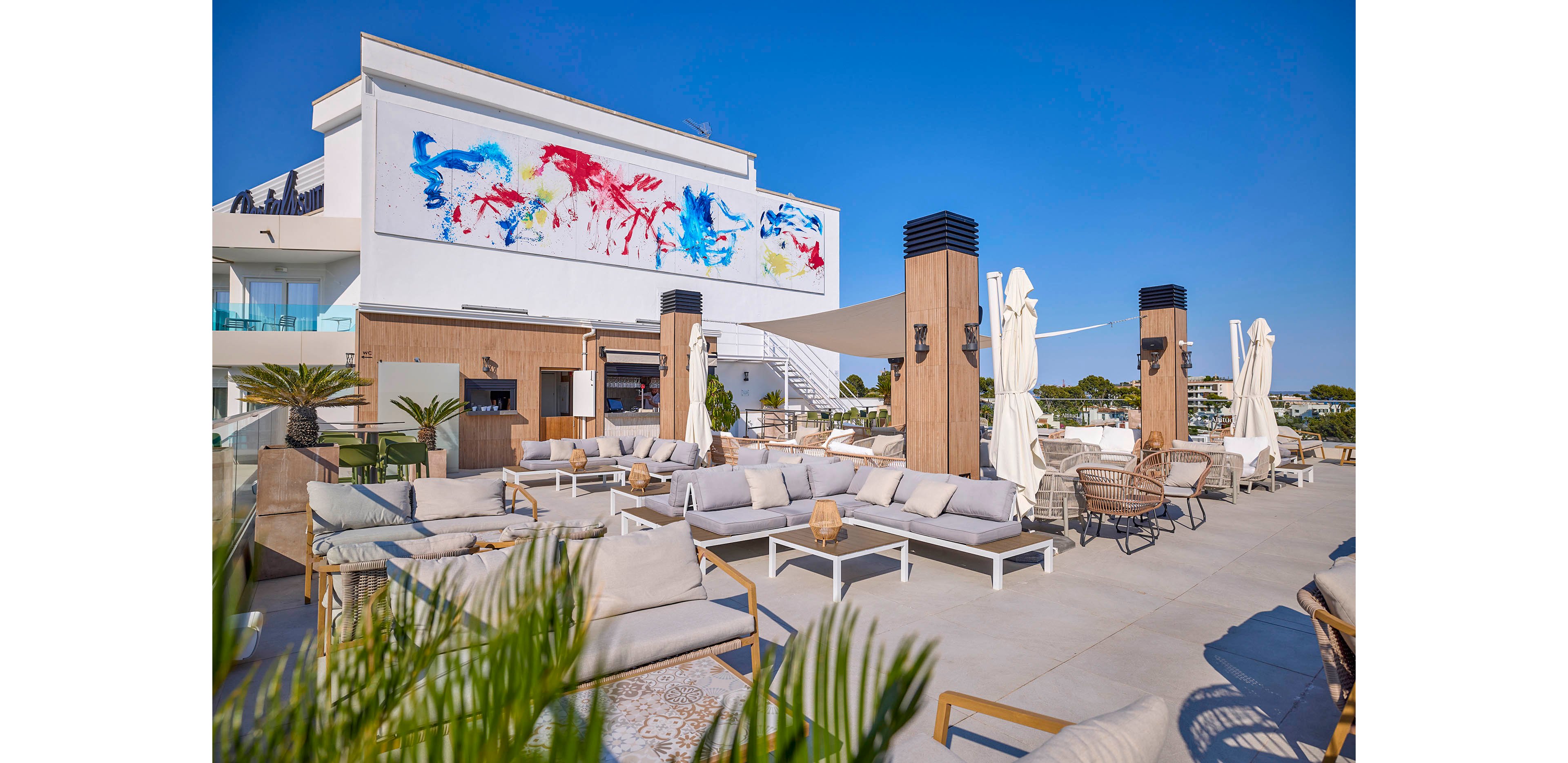 A contemporary rooftop lounge features cushioned outdoor sofas and low tables arranged for relaxed socializing. Wooden pillars, white umbrellas, and potted greenery create a stylish open-air setting under a clear blue sky. A large abstract mural with bold red, blue, and yellow brushstrokes decorates the building facade, adding vibrant color to the minimalist architecture. The scene suggests a sunny, upscale leisure environment ideal for gatherings and relaxation.