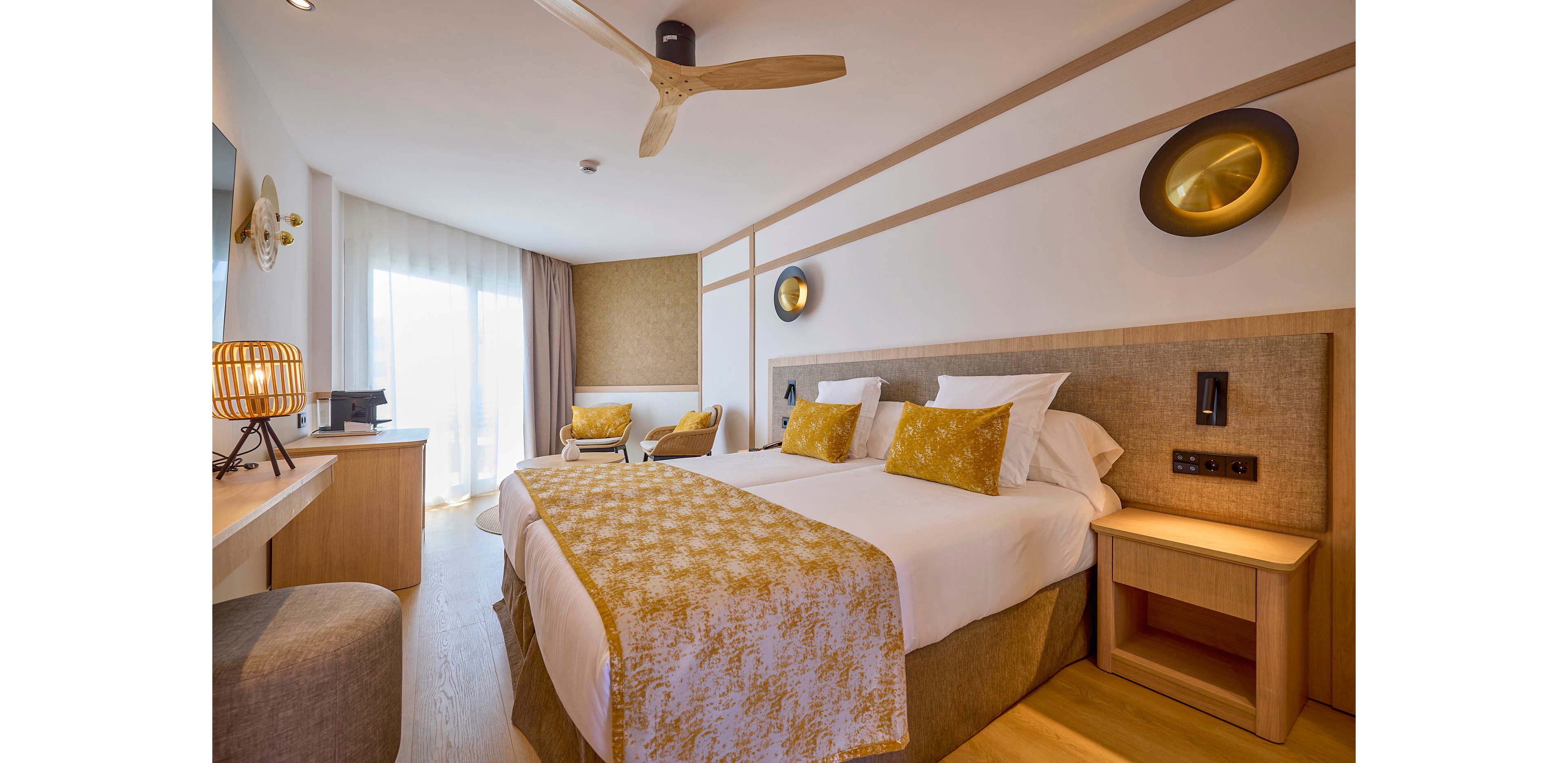 Contemporary hotel bedroom featuring a large double bed with white linens and patterned yellow throw pillows. The room is decorated in warm neutral tones with wooden furniture, including a bedside table, desk, and cushioned stool. Natural light streams in through sheer curtains covering a large window or balcony door. Decorative wall lights, a ceiling fan, and a woven lamp add cozy ambiance to the clean, minimalist interior.