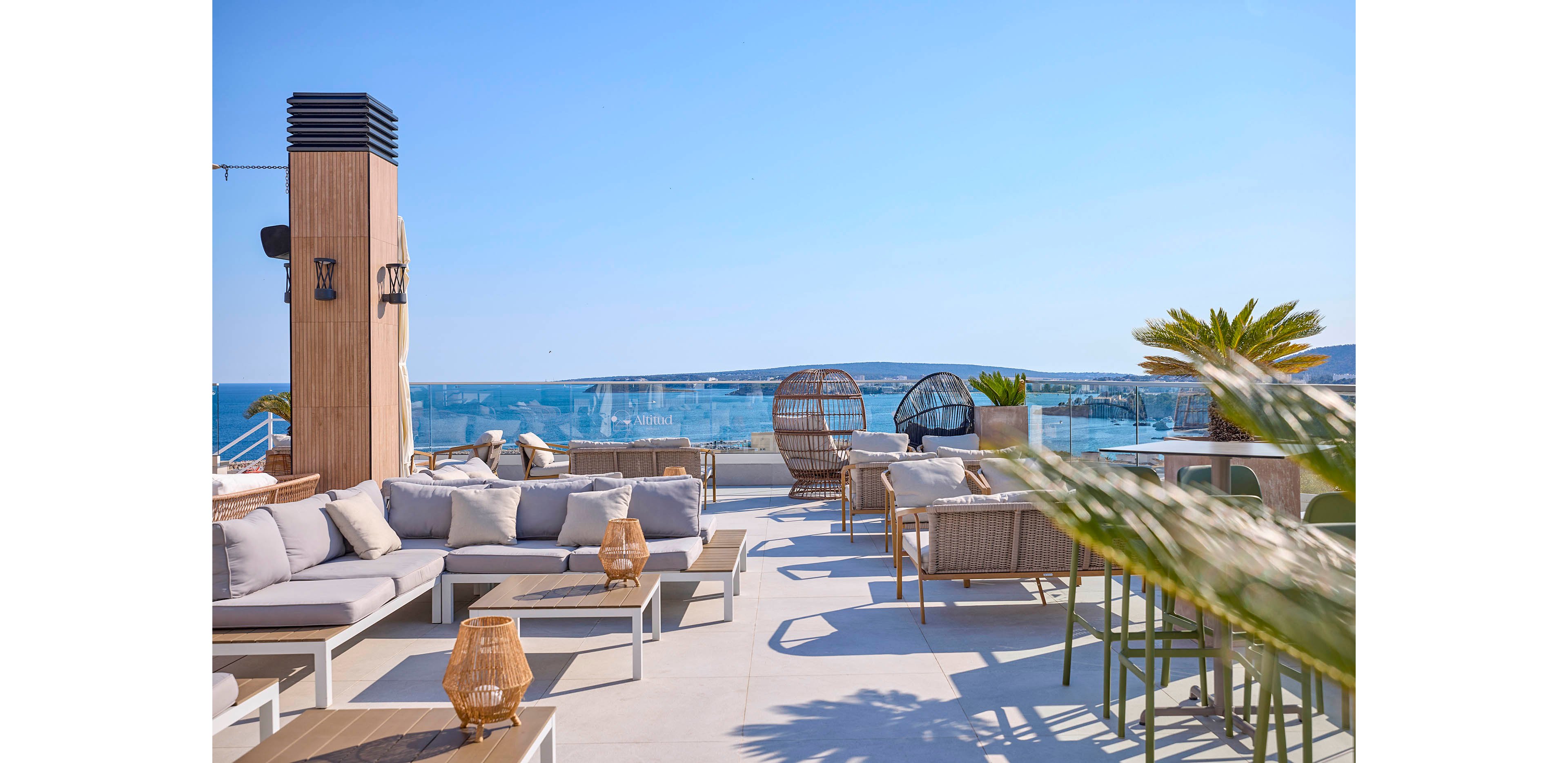 A contemporary rooftop terrace features cushioned sofas and wicker chairs arranged around low tables. Decorative lanterns and potted plants add warmth to the open-air seating area. Glass railings reveal a panoramic view of the sea and distant coastline under a clear blue sky. The scene suggests a relaxed, upscale outdoor social space bathed in bright daylight.