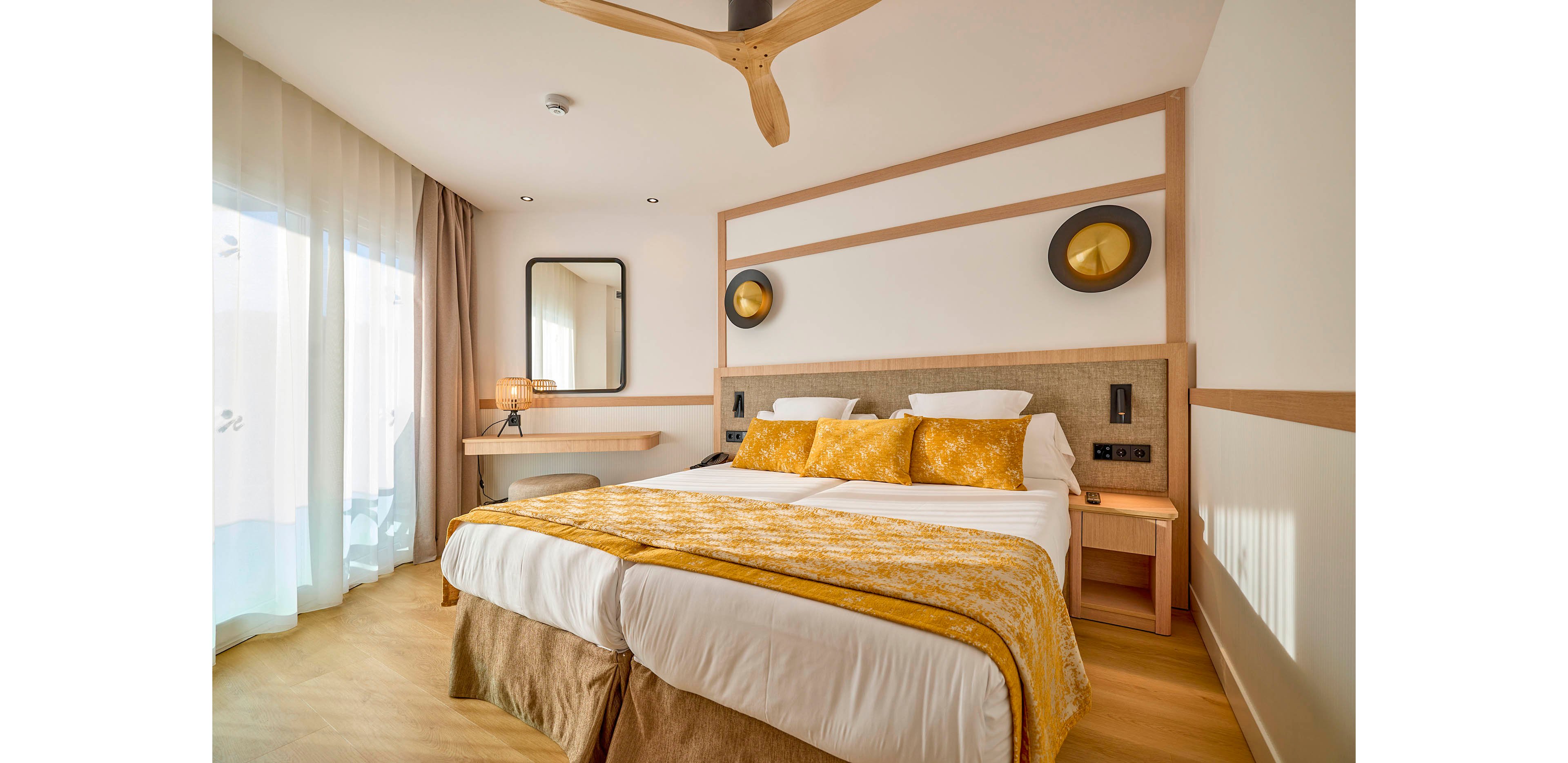 A contemporary hotel room features a large double bed with white linens and yellow accent pillows and throw. The space is warmly lit, with natural light streaming through sheer curtains covering a glass door. Minimalist wooden furniture includes bedside tables, a desk with a chair, and a wall-mounted mirror. Neutral tones and clean lines create a cozy yet modern atmosphere suitable for relaxation.