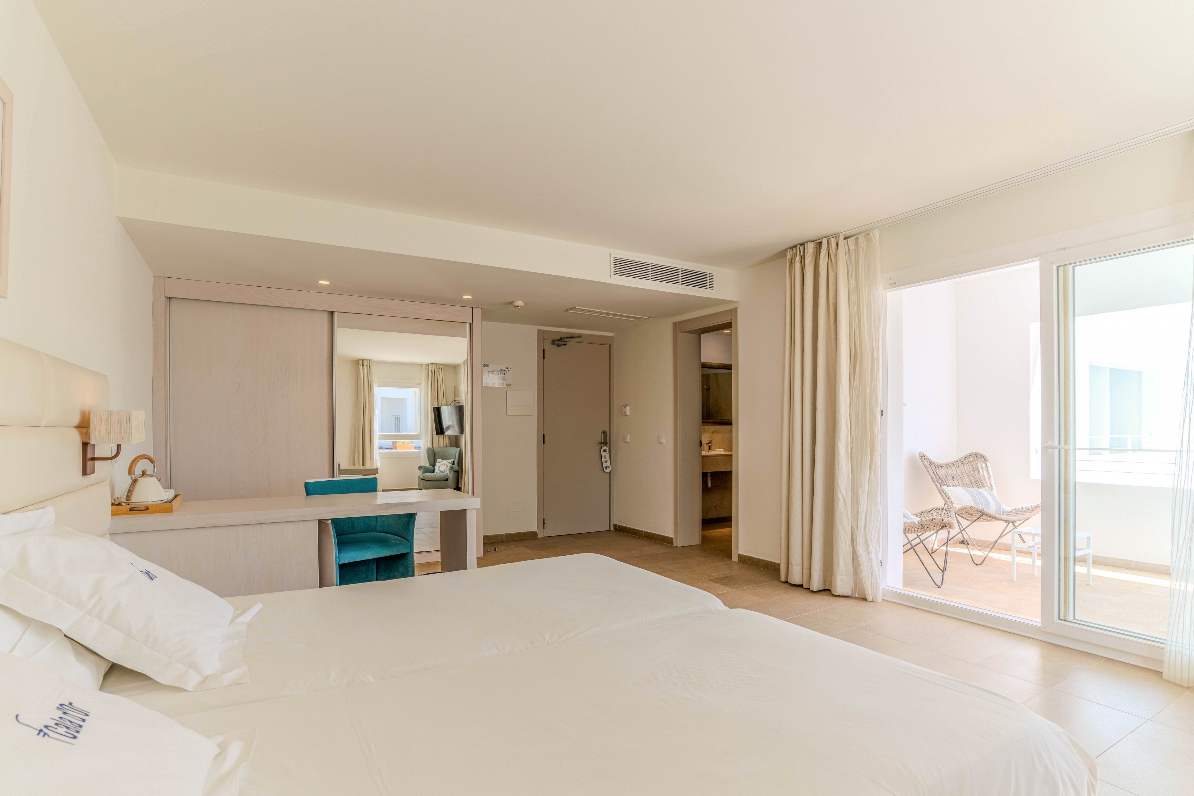 A bright, modern hotel room features a neatly made double bed with white linens in the foreground. A desk with a turquoise chair and a large mirror sits against the wall near the entrance door. Floor-to-ceiling curtains open onto a balcony with outdoor seating, allowing natural light to fill the space. Neutral tones and minimal decor create a clean, relaxing atmosphere suitable for vacation or business travel.