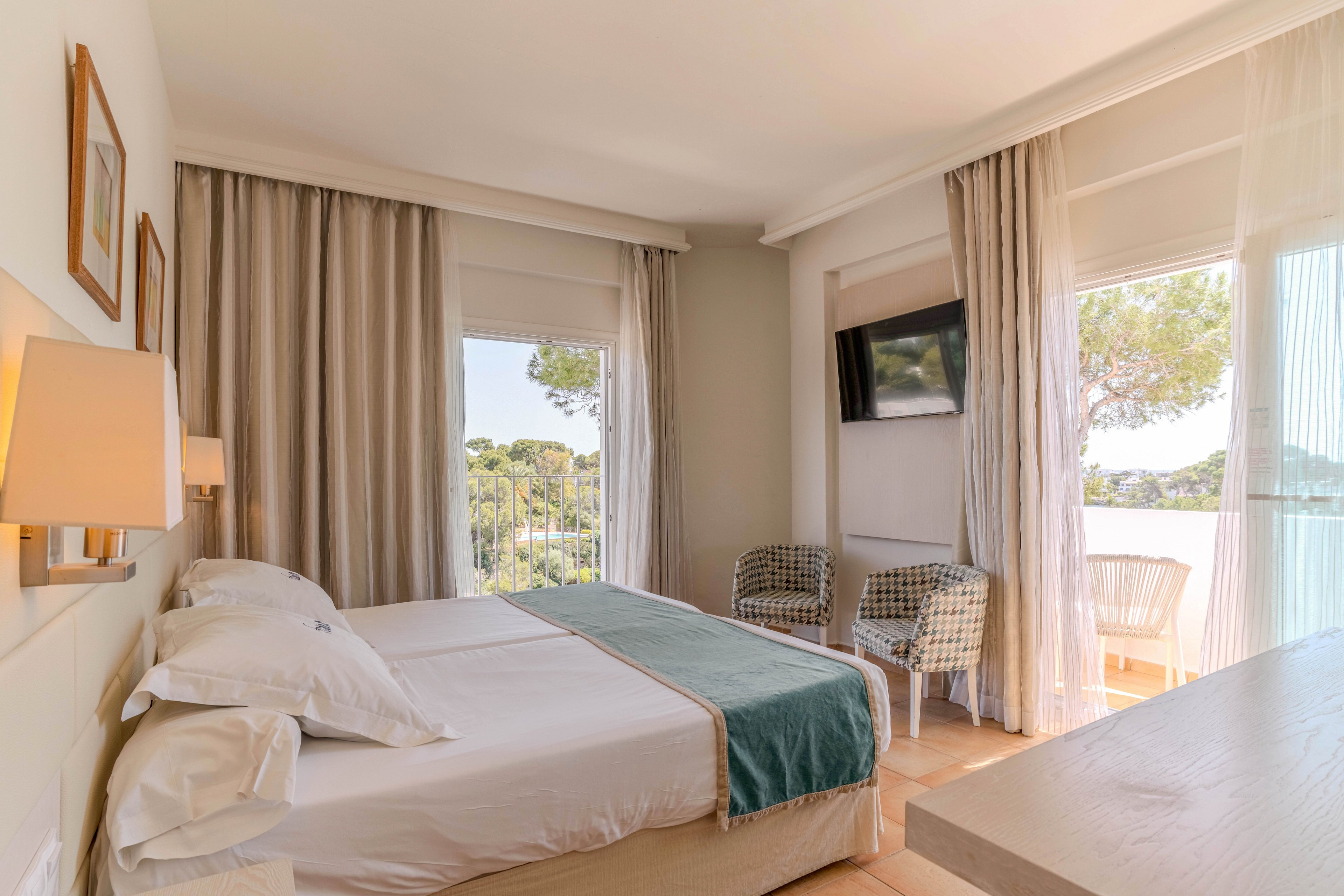 A bright, modern hotel room features a neatly made double bed with white linens and a teal runner. Large windows with sheer curtains let in abundant natural light and offer views of trees outside. Two woven armchairs and a wall-mounted television create a comfortable seating area near the balcony. Soft neutral tones and minimalist decor give the space a calm, airy atmosphere.