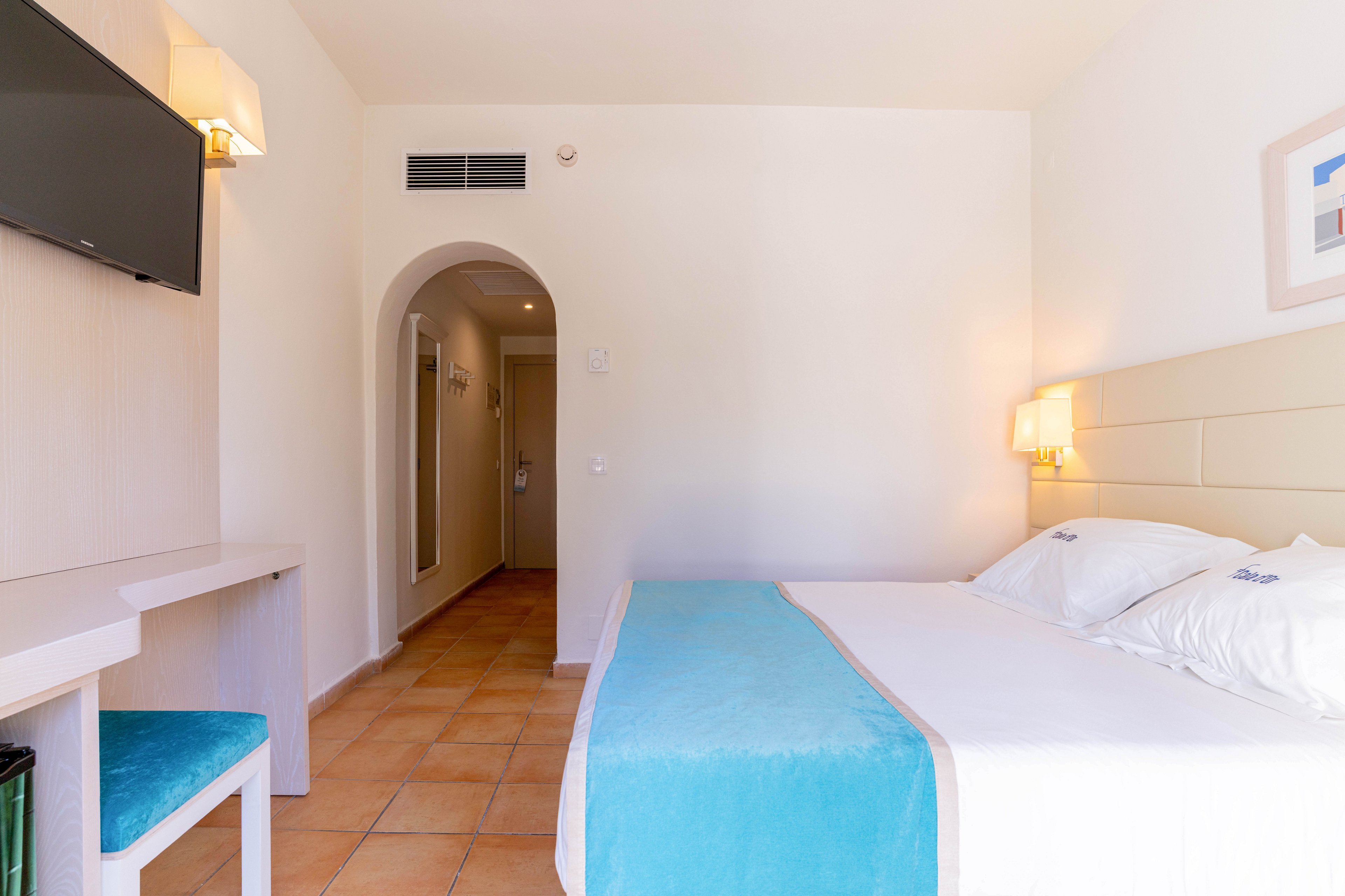 A bright, minimalist hotel room features a neatly made twin or double bed with white linens and a turquoise bed runner. The space includes a simple desk with a matching turquoise stool and a wall-mounted flat-screen television. Warm terracotta floor tiles lead toward an arched doorway and a hallway with additional doors. Soft wall sconces and neutral walls create a clean, inviting atmosphere suitable for vacation or business travel.