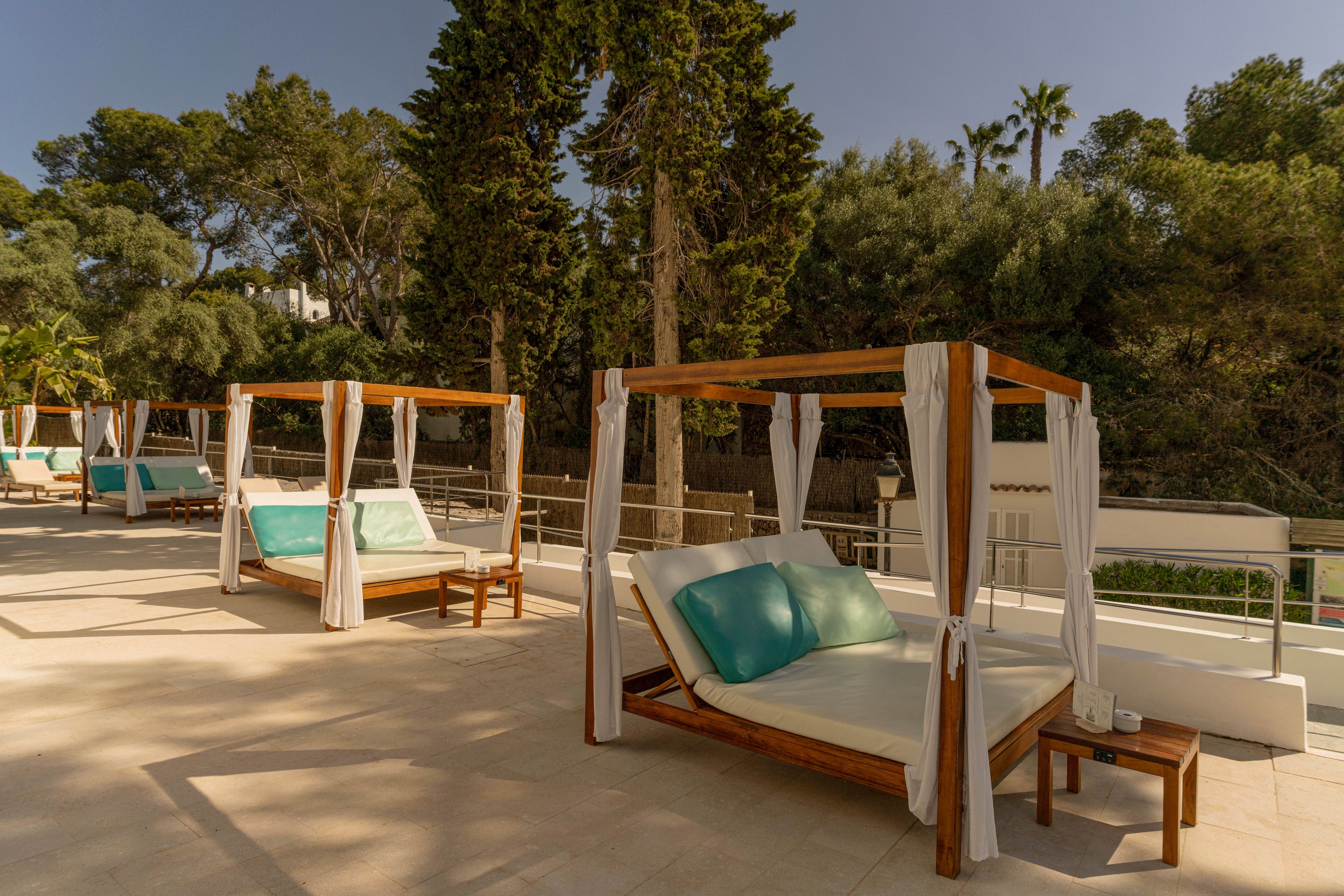Several wooden cabana-style daybeds with white curtains and teal cushions are arranged on a spacious outdoor terrace. The scene is sunlit, with soft shadows cast across the light stone flooring. Tall trees and lush greenery form a natural backdrop behind the terrace, suggesting a resort or spa setting. Small side tables beside the beds hold glasses and carafes, enhancing the relaxed, luxurious atmosphere.