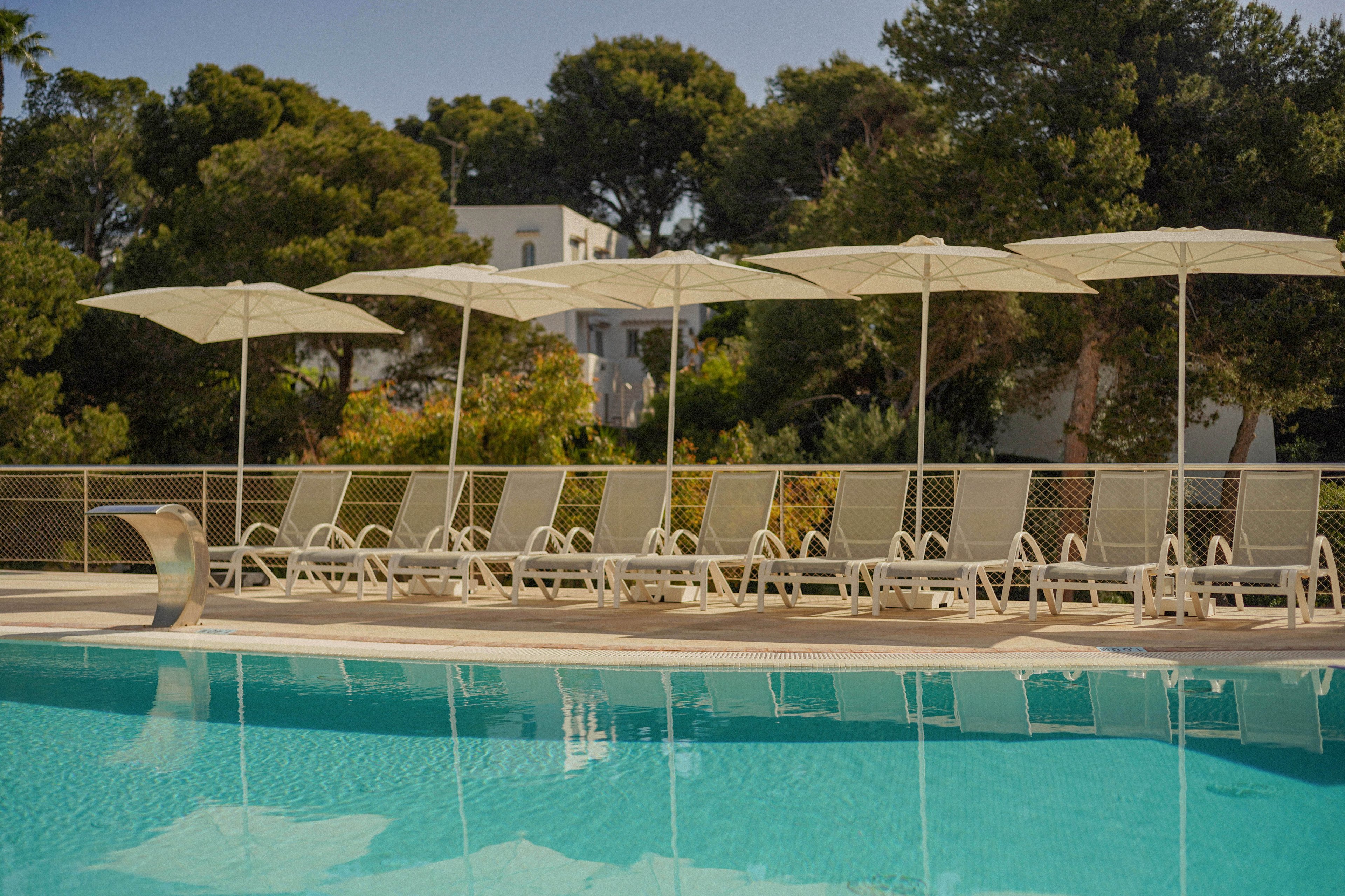 A tranquil outdoor swimming pool reflects the clear blue water in the foreground. A row of empty sun loungers sits neatly arranged under large white umbrellas along the pool deck. Trees and greenery frame the scene, with modern white buildings visible in the background. The overall atmosphere feels calm, sunny, and inviting, suggesting a quiet resort or hotel setting.