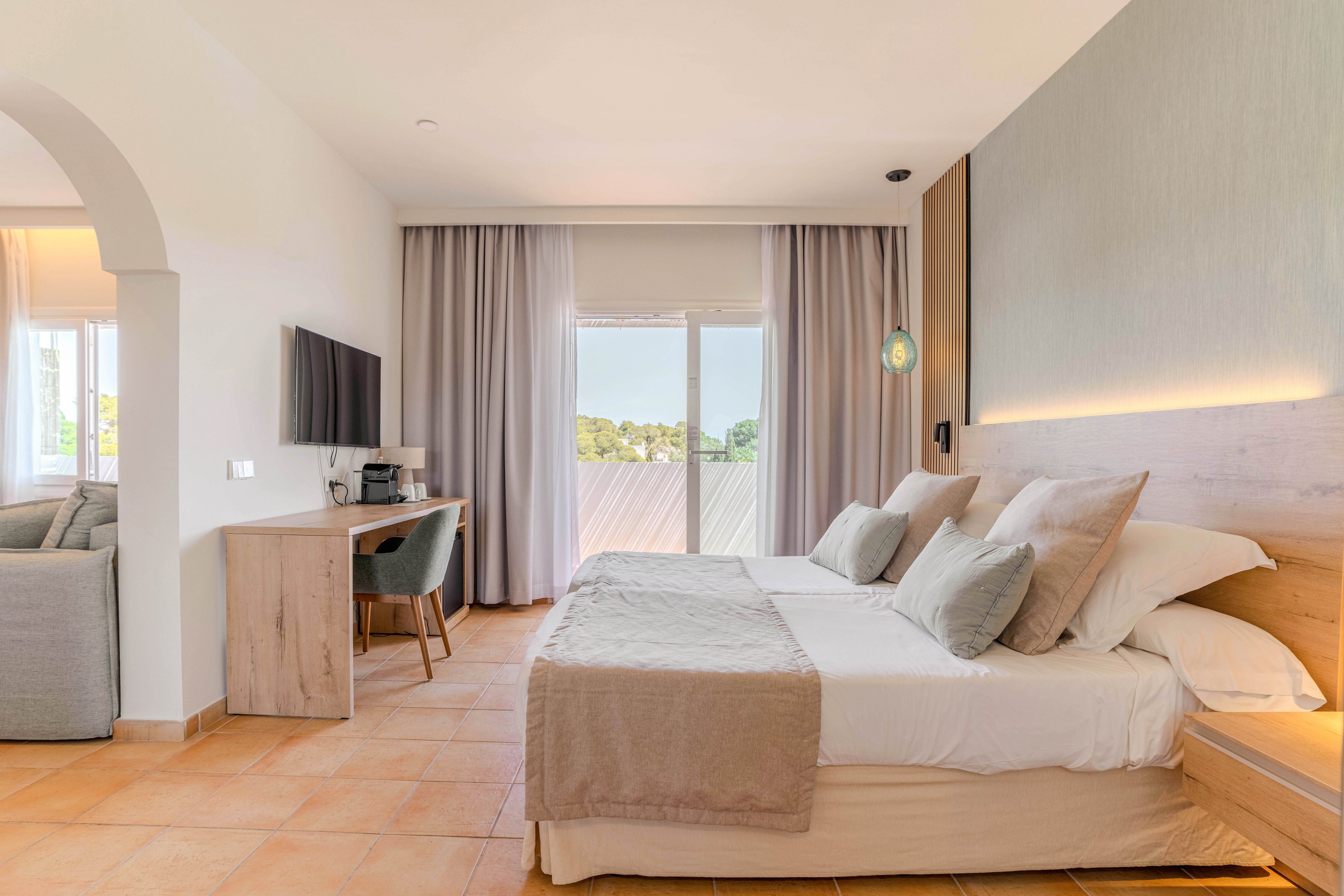 A contemporary hotel bedroom features a large double bed with neutral bedding and multiple pillows. A wall-mounted flat-screen TV and simple wooden desk with chair sit opposite the bed, creating a functional workspace. Floor-to-ceiling curtains frame glass doors that open to a balcony with a scenic outdoor view. Warm tile flooring and soft ambient lighting contribute to a calm, inviting atmosphere.