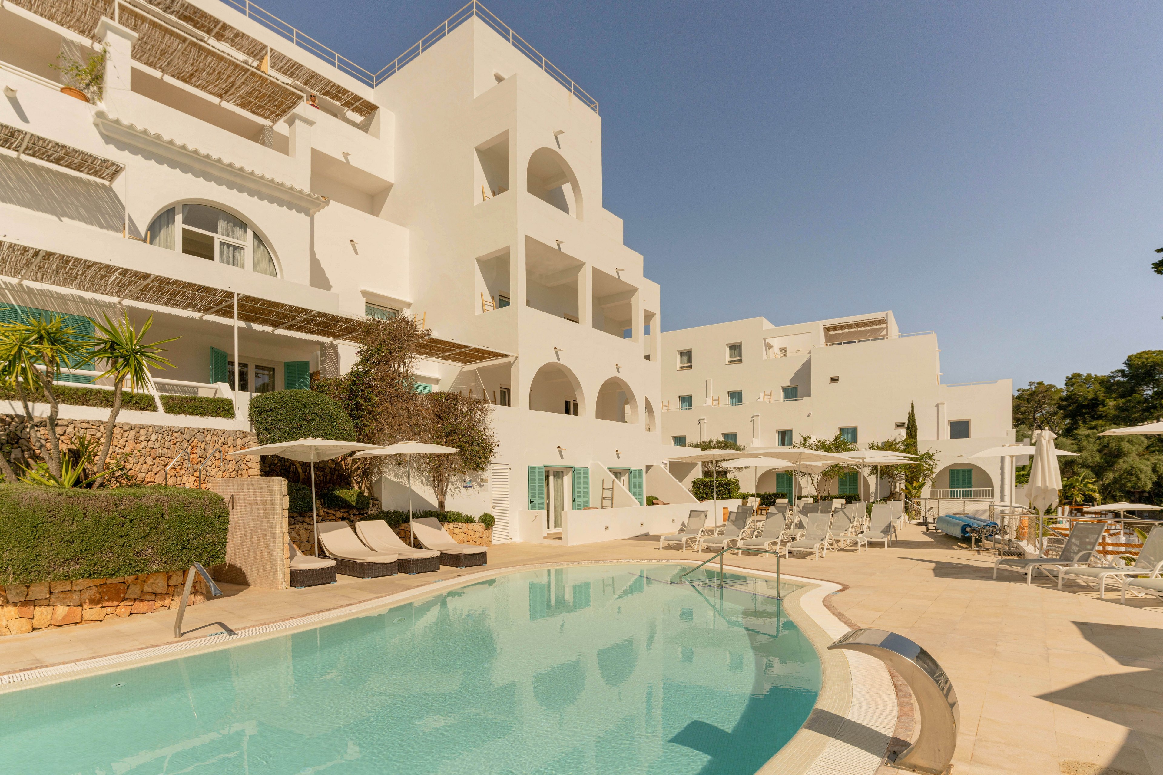 A bright white resort complex features multiple levels of balconies and arched windows overlooking a clear outdoor swimming pool. Sun loungers and umbrellas line the poolside terrace, suggesting a relaxing vacation setting. The architecture has a Mediterranean feel, with clean geometric shapes and light-colored surfaces reflecting the warm sunlight. Lush greenery and potted plants add contrast to the pale buildings and blue water.
