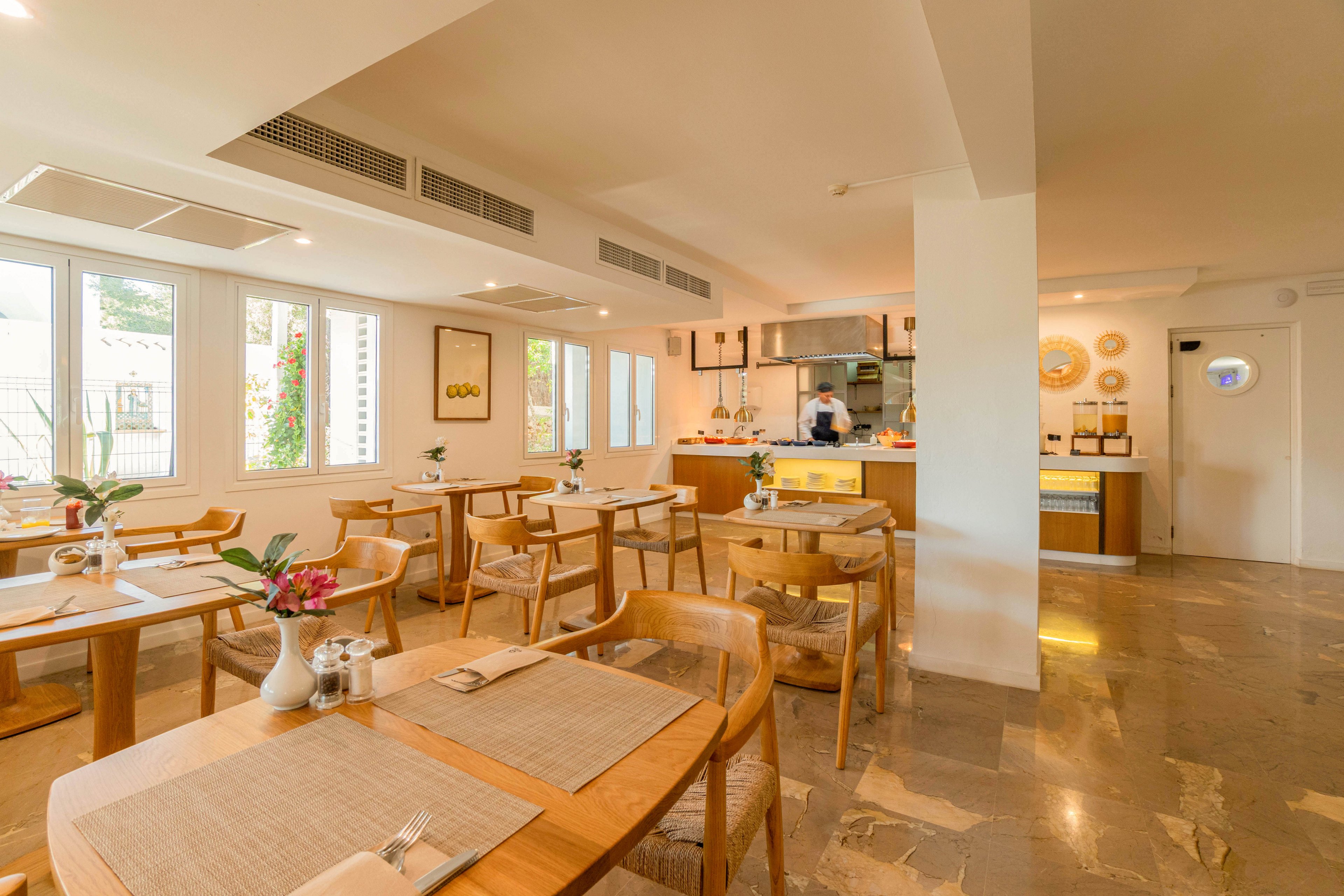 A bright, modern restaurant dining area features wooden tables and chairs neatly arranged with placemats and simple floral centerpieces. Large windows on one side allow natural light to fill the space, creating an airy and welcoming atmosphere. In the background, an open kitchen or service counter is visible, where a staff member prepares or serves food. Warm lighting, neutral tones, and clean lines contribute to a contemporary yet cozy dining environment.