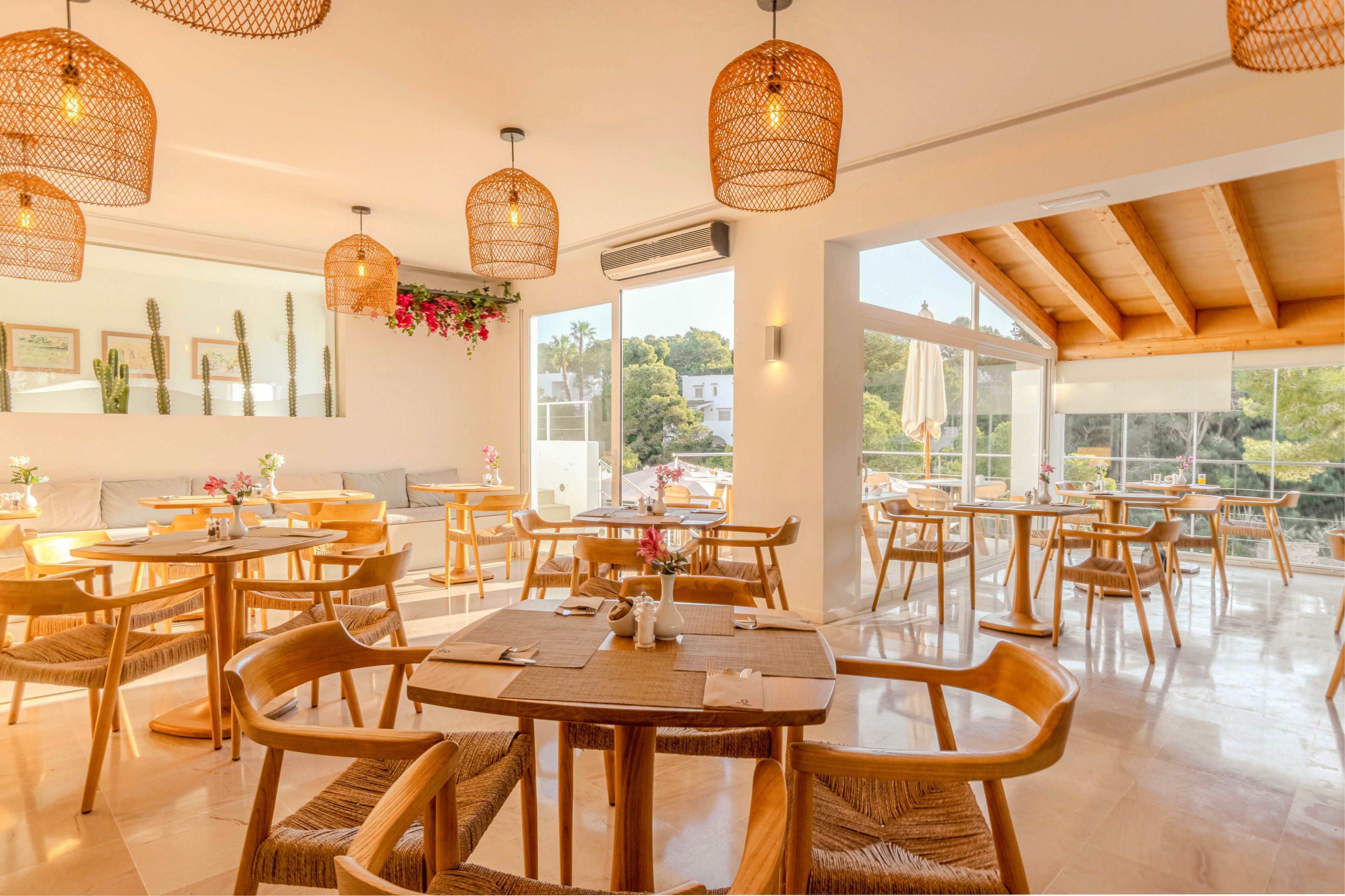 A sunlit restaurant dining area features round wooden tables and matching chairs arranged neatly across the room. Large windows and glass doors let in abundant natural light and offer views of greenery outside. Woven pendant lights hang from the ceiling, casting a warm glow over the neutral-toned space. Decorative plants, flowers, and wall art add a relaxed, contemporary Mediterranean feel to the interior.