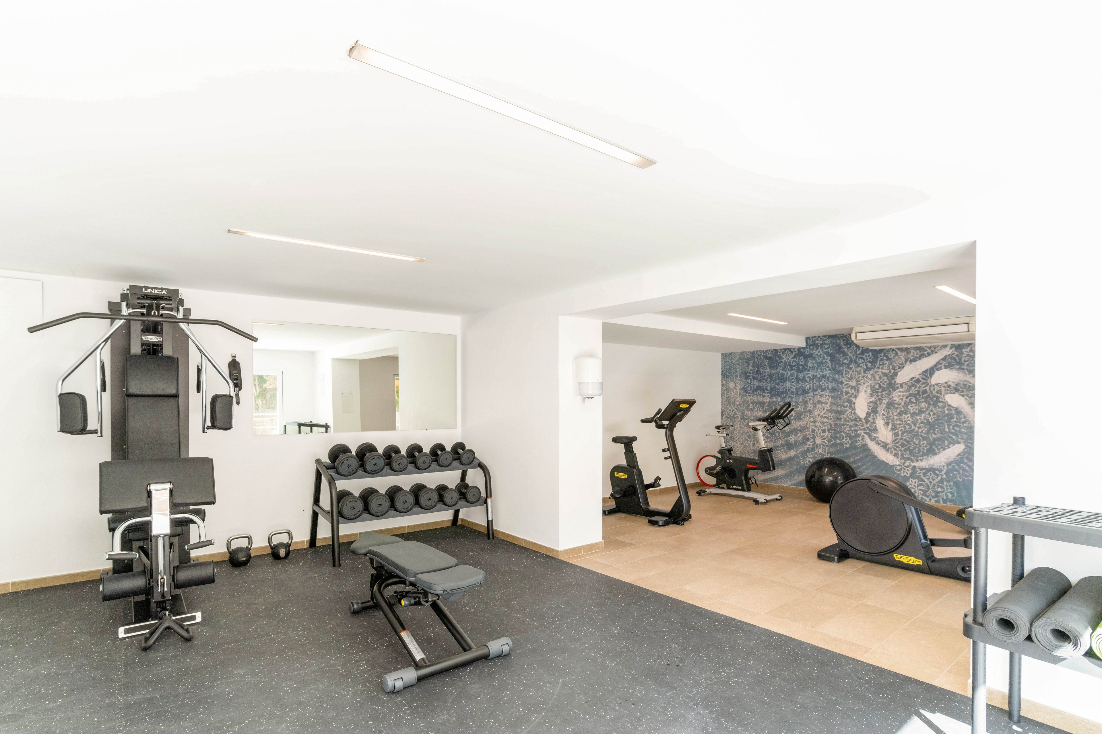 A bright, modern home gym features a variety of fitness equipment neatly arranged in a clean space. In the foreground, a weight bench, multi-gym machine, and a rack of dumbbells sit on a dark rubber floor. The background area includes stationary bikes, an exercise ball, and additional machines on a light tiled floor. White walls and a large abstract mural add to the airy, minimalist atmosphere.