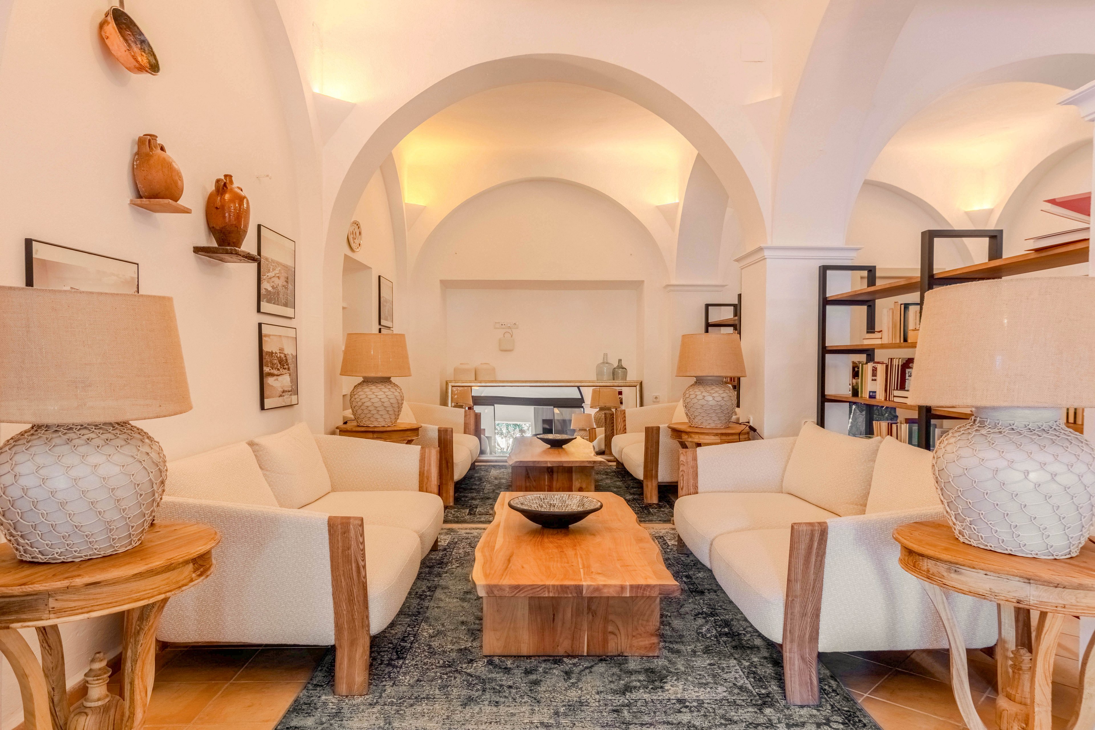 A cozy living room features two cream-colored sofas facing each other around a solid wooden coffee table. Large textured table lamps sit on matching side tables, casting a warm glow across the space. The room is framed by elegant white arches, with shelves of books and decorative pottery adding character. Soft lighting and neutral tones create a calm, inviting atmosphere ideal for relaxation or conversation.