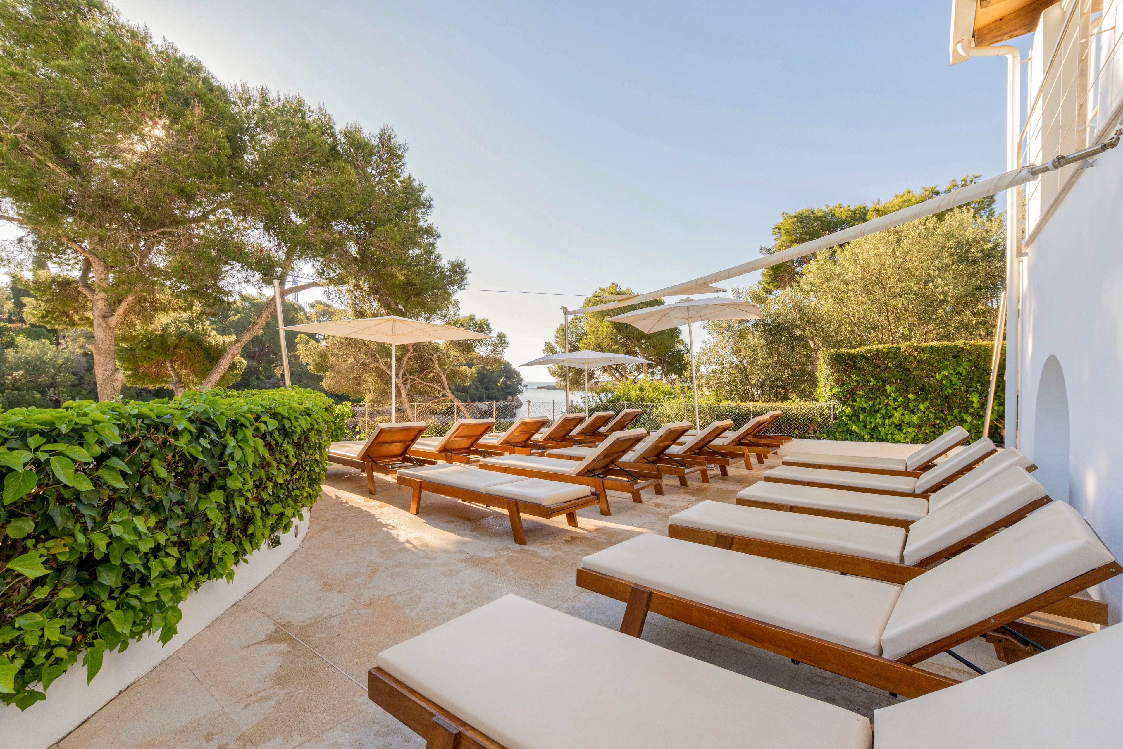 A row of cushioned wooden lounge chairs lines a bright terrace overlooking the water. Large white umbrellas provide shade, while lush green hedges and tall trees frame the outdoor space. The scene suggests a tranquil coastal resort or villa, with clear skies and soft natural light. The overall atmosphere is relaxed, inviting guests to sunbathe or rest by the seaside.