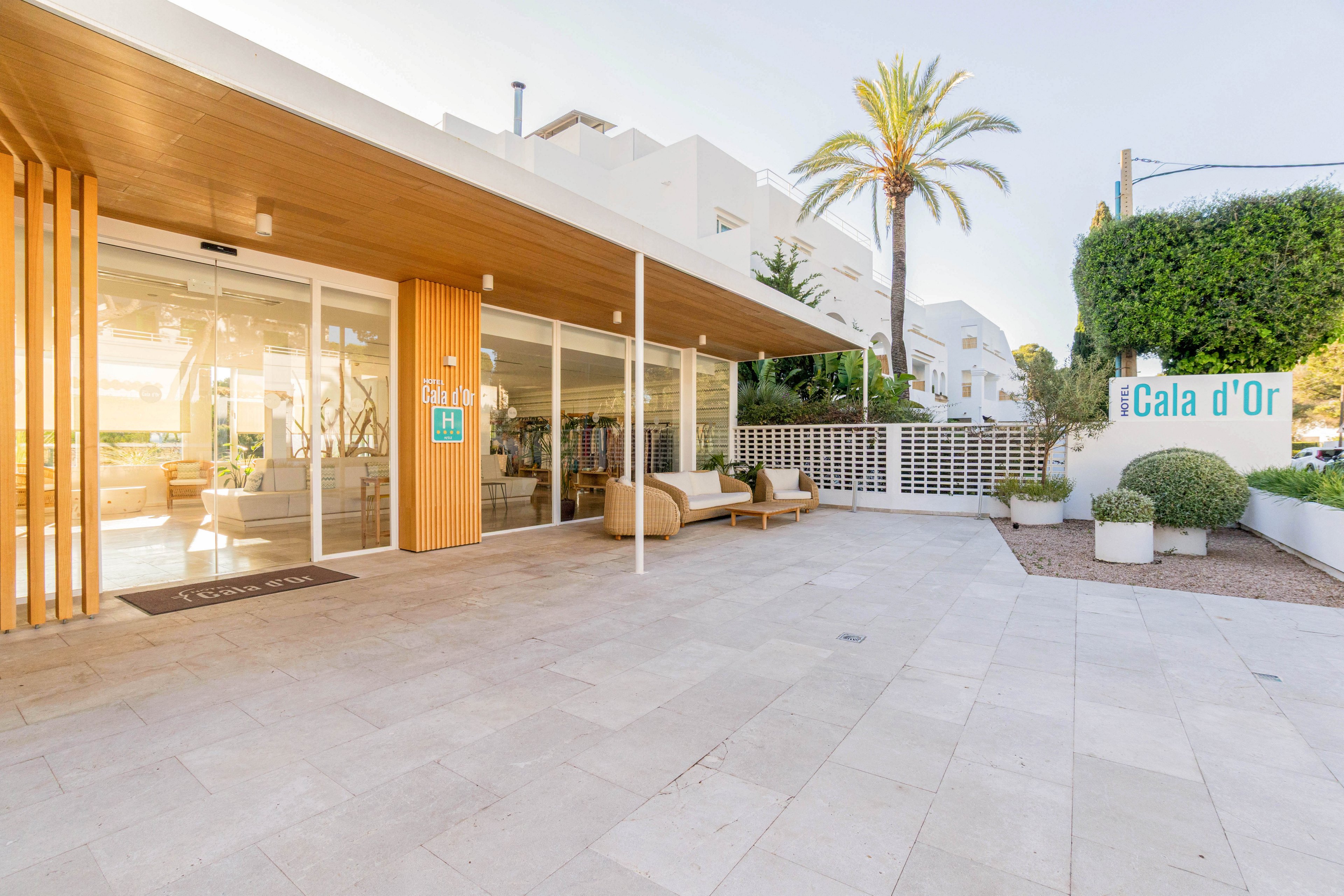 A modern boutique or reception area with large glass doors opens onto a spacious tiled courtyard. Warm wooden accents and minimalist outdoor seating create a welcoming, contemporary look. A sign reading "Cala d'Or" stands near manicured greenery and a tall palm tree. Bright daylight and clean white architecture suggest a relaxed Mediterranean coastal setting.