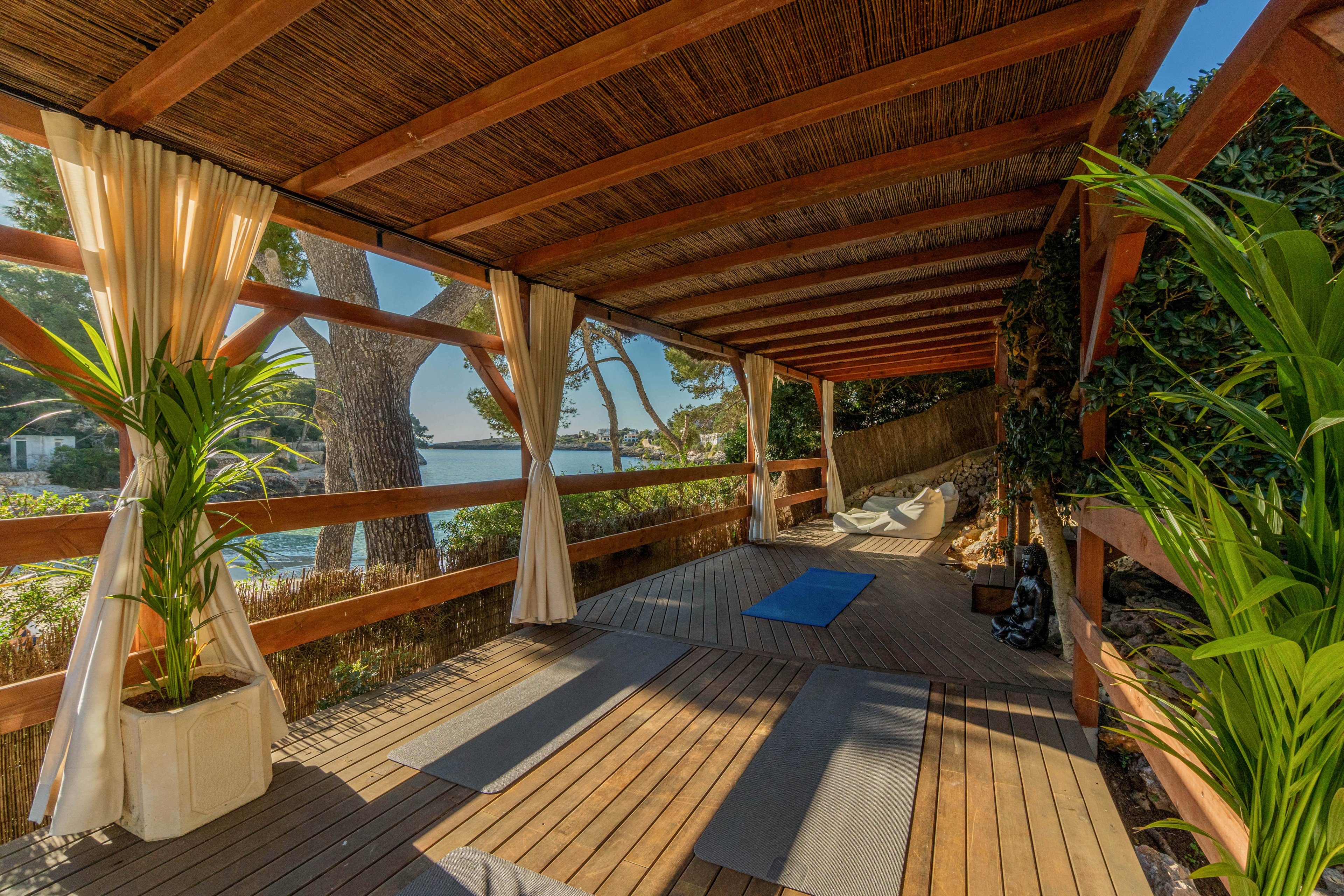 An open-air wooden deck is set up as a yoga space with several mats laid out on the floor. The structure is covered with a slatted roof and framed by light curtains tied to wooden posts. Lush green plants in pots decorate the corners, and sunlight filters through the trees and railing. In the background, a calm coastal landscape with water and trees creates a serene atmosphere. The scene suggests a peaceful retreat for outdoor exercise and relaxation.