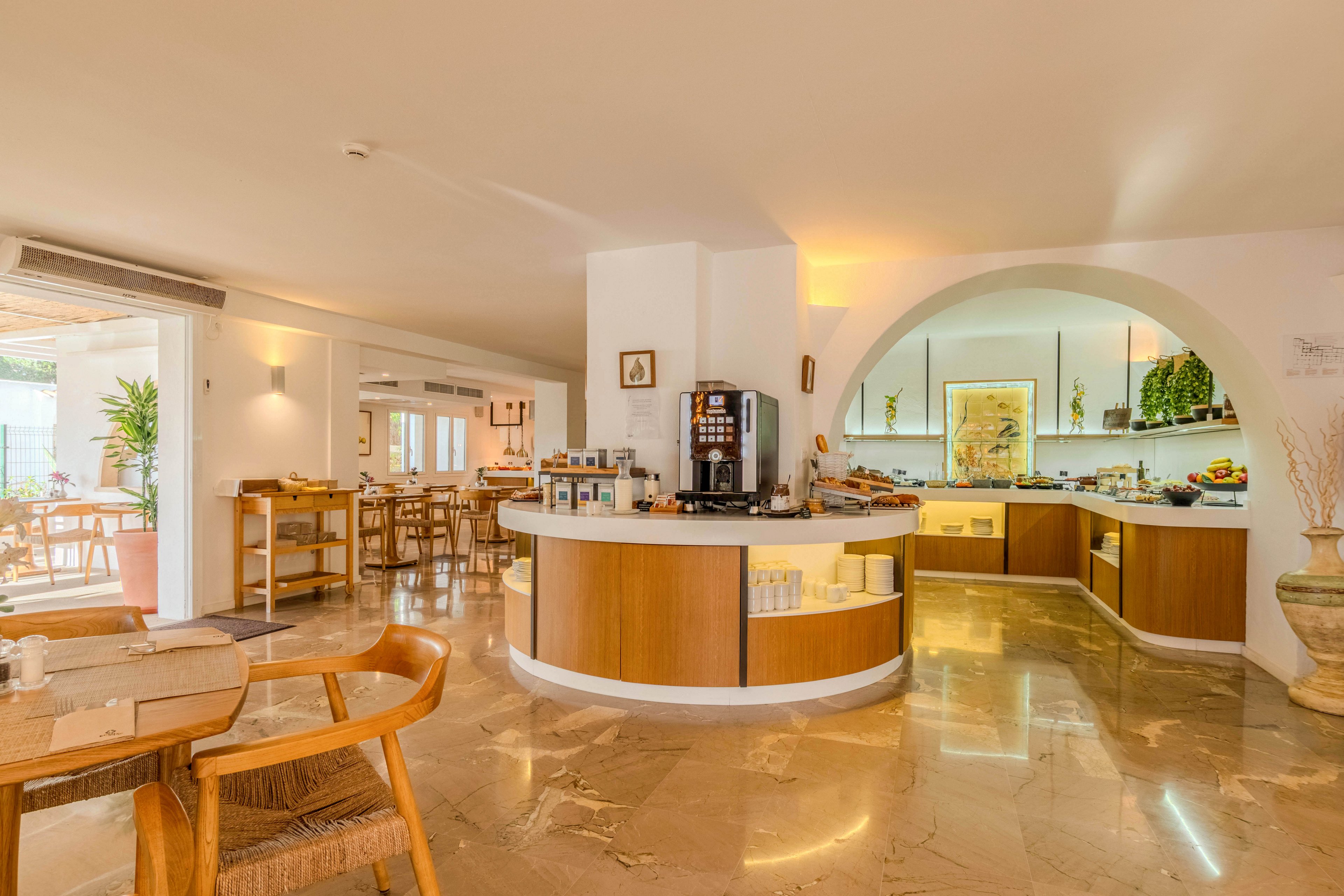 Spacious hotel dining room featuring a central breakfast buffet counter with coffee machines and food displays. Warm wooden furniture and marble floors create an inviting, modern atmosphere. Large windows and open doors let in natural light, connecting the interior with an outdoor seating area. The space appears clean, organized, and ready for guests to enjoy a relaxed meal.