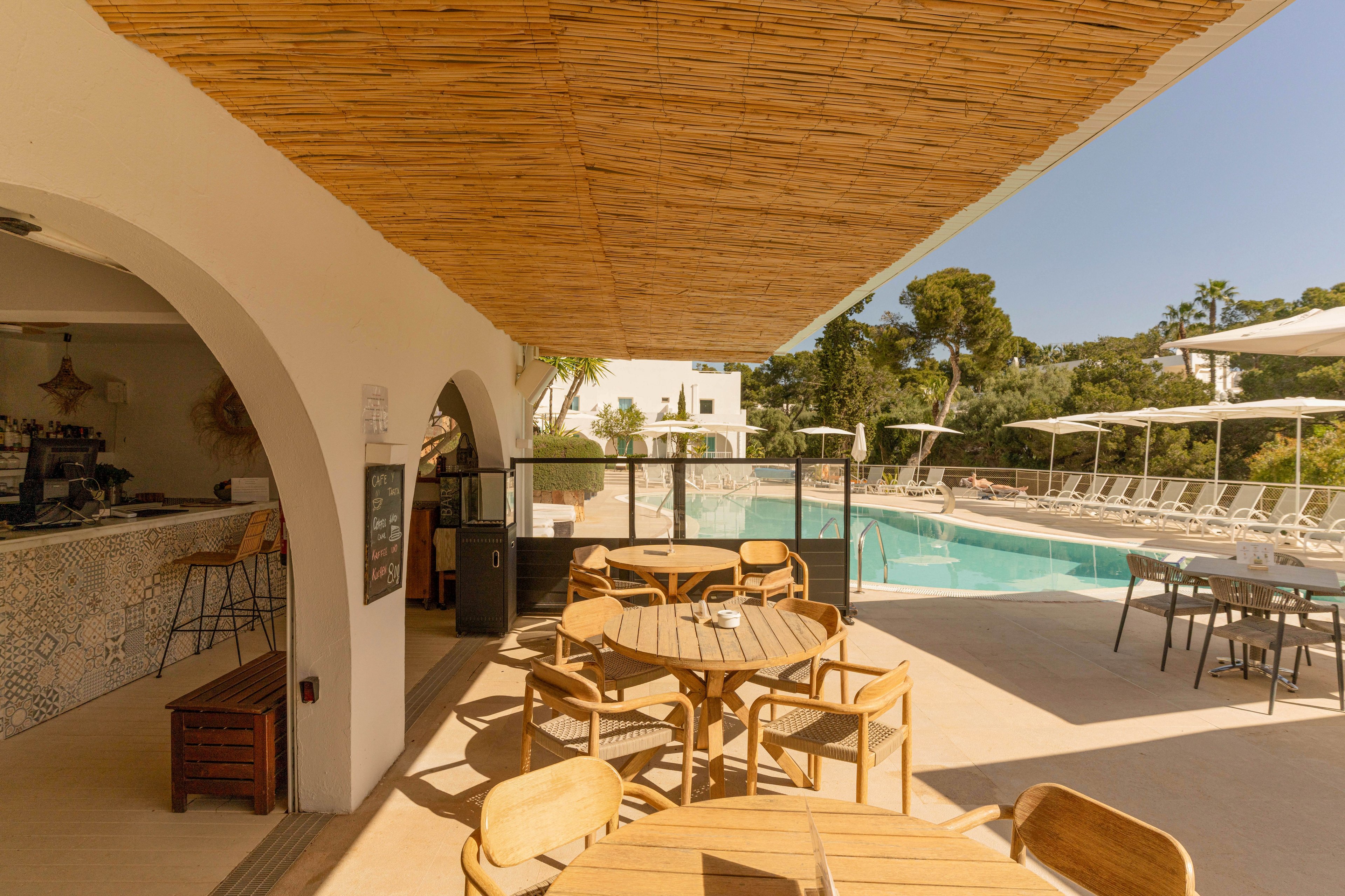 An inviting outdoor café terrace overlooks a bright swimming pool on a sunny day. Wooden tables and chairs sit beneath a woven shade roof, creating a relaxed seating area. The bar area with tiled walls and stools is tucked under white arches to the left. Sun loungers and umbrellas line the poolside, surrounded by greenery and trees in the background.
