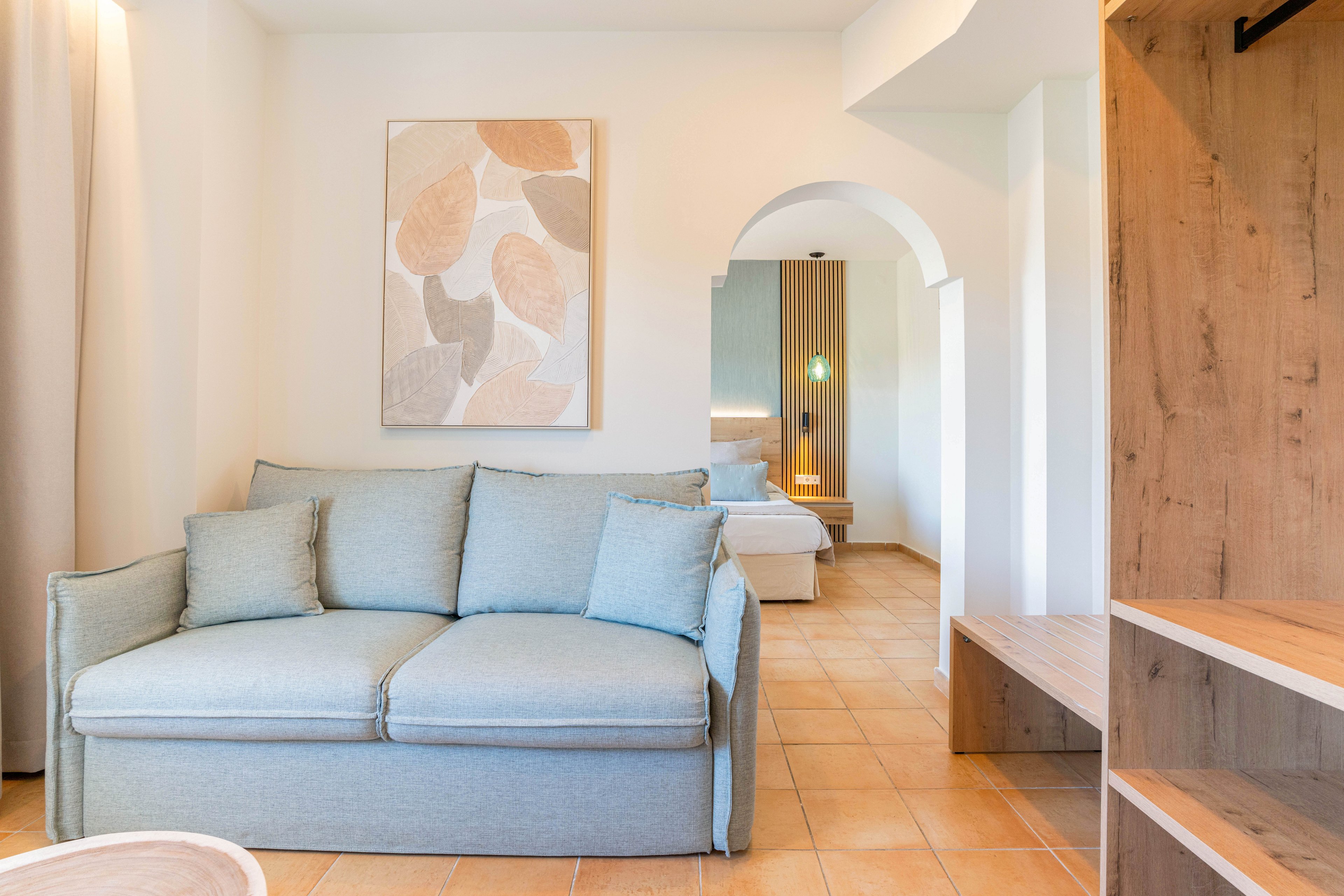 A light blue upholstered sofa sits in a bright, modern room with neutral walls and tiled flooring. An abstract leaf artwork hangs above the sofa, adding a soft decorative touch. Through an arched doorway, a neatly made bed and warm bedside lighting are visible, suggesting a connected bedroom area. Wooden shelving and built-in furniture complete the minimalist, contemporary interior design.