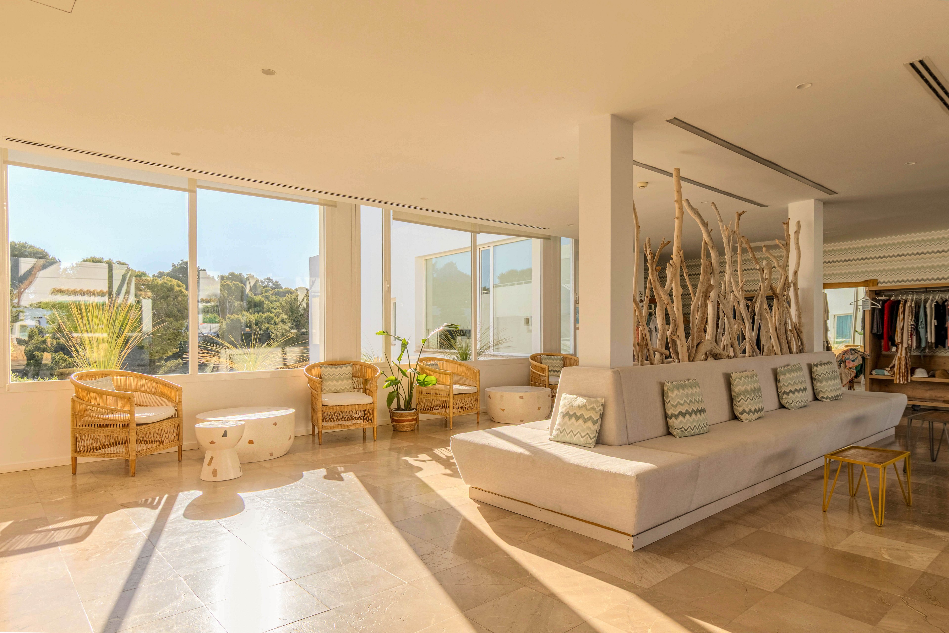 A spacious sunlit lounge features a long beige sectional sofa accented with patterned cushions. Large floor-to-ceiling windows reveal a view of trees and nearby buildings, filling the room with natural light. Wicker chairs, small side tables, and decorative driftwood elements create a relaxed coastal atmosphere. In the background, open shelving with hanging clothes suggests a boutique or resort setting.