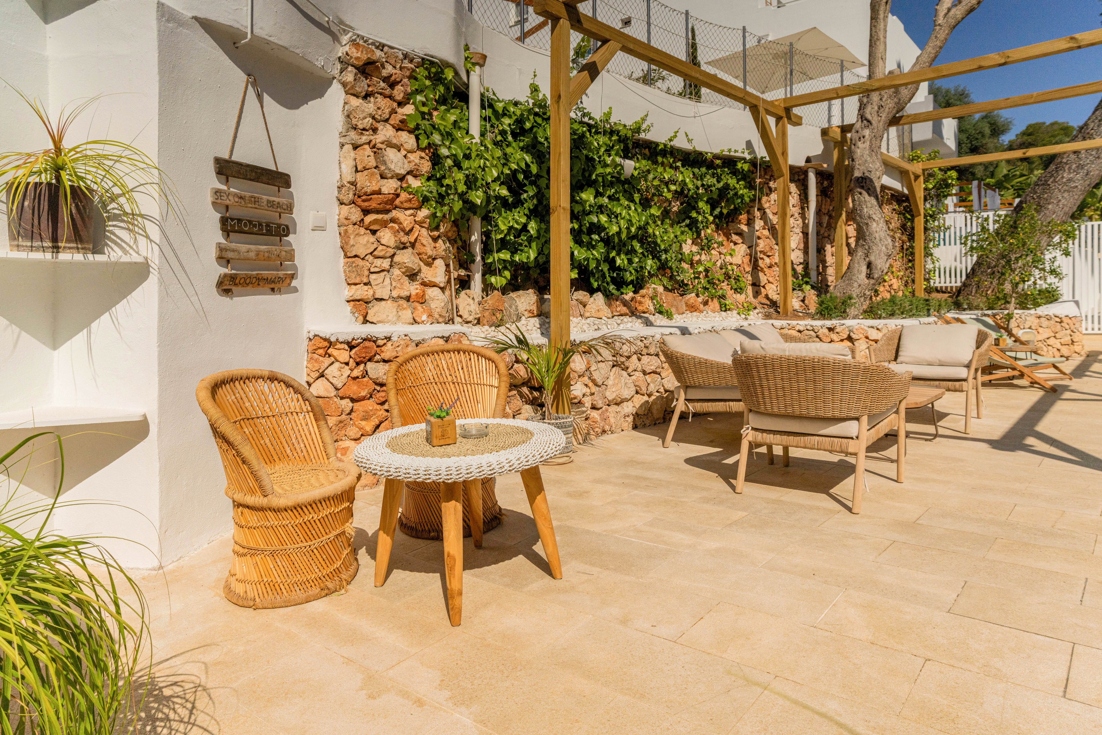 An inviting outdoor patio features wicker chairs and a small round table set against a sunlit stone wall. A pergola with climbing greenery provides partial shade over additional lounge seating in the background. The warm beige floor tiles and white walls create a bright Mediterranean feel. Potted plants and decorative wall shelves add natural accents to the cozy terrace.