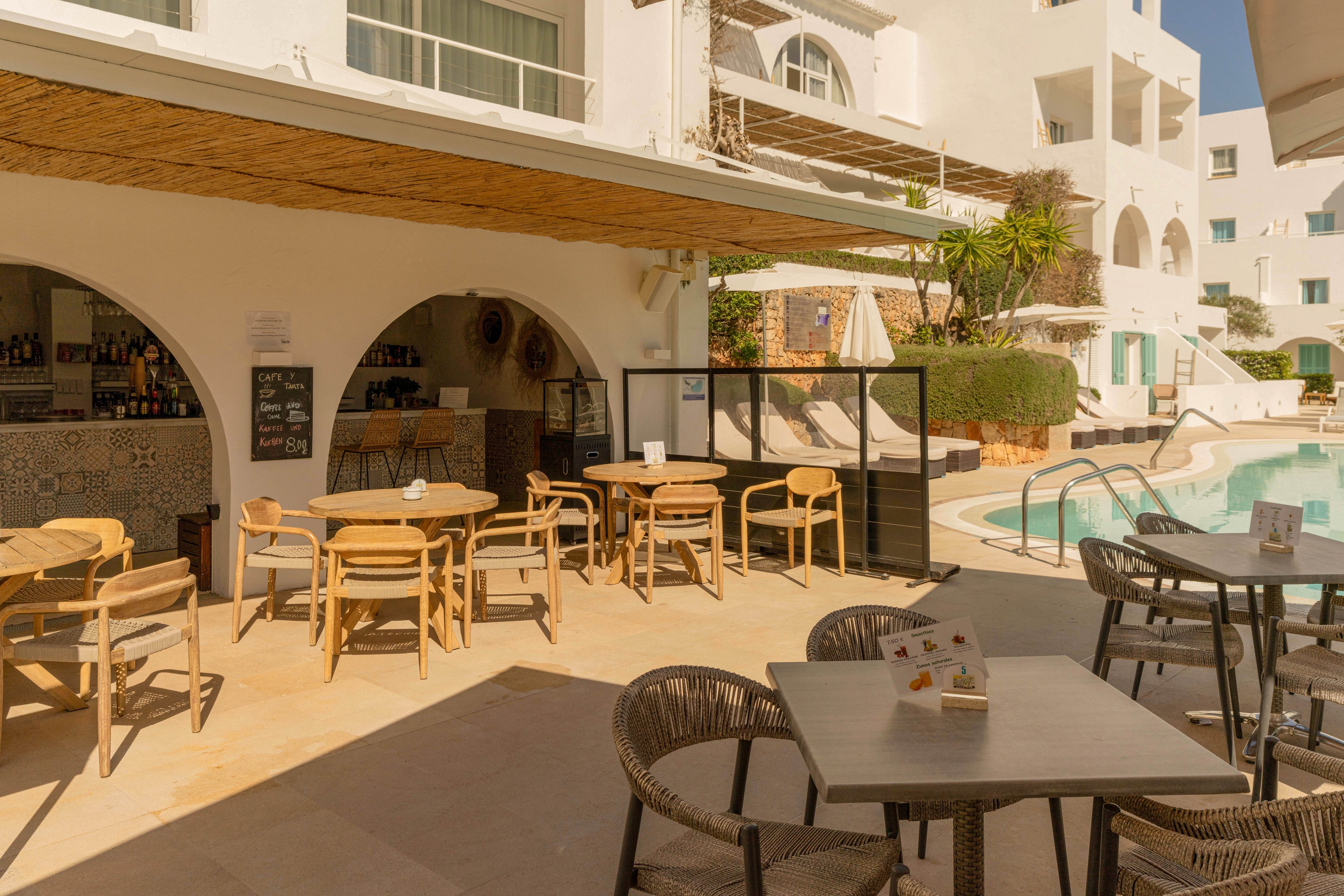 An outdoor café terrace sits beside a bright swimming pool at a white Mediterranean-style resort. Wooden tables and chairs are arranged in the sun and shade, with menus placed on several tables. The bar area is tucked under an arched canopy, displaying bottles and a chalkboard menu on the wall. White apartment-style buildings with balconies and windows surround the pool area, creating a relaxed holiday atmosphere. Warm sunlight and soft shadows give the scene a calm, summery feel.