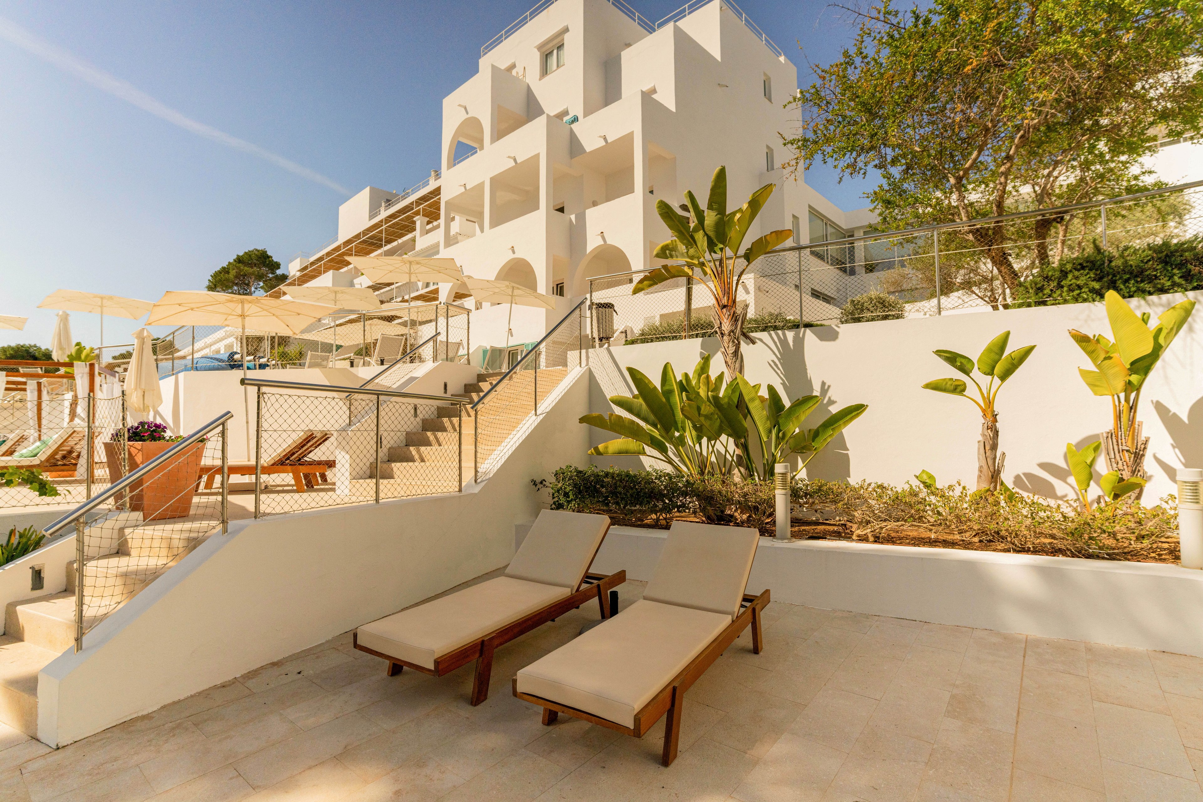 A modern white Mediterranean-style hotel is shown with multiple levels and arched balconies. In the foreground, two cushioned wooden sun loungers sit on a tiled terrace near landscaped greenery. Stairs lead up to an elevated area with umbrellas and additional seating, suggesting a resort or vacation setting. Warm sunlight and clear skies create a relaxed, summery atmosphere.