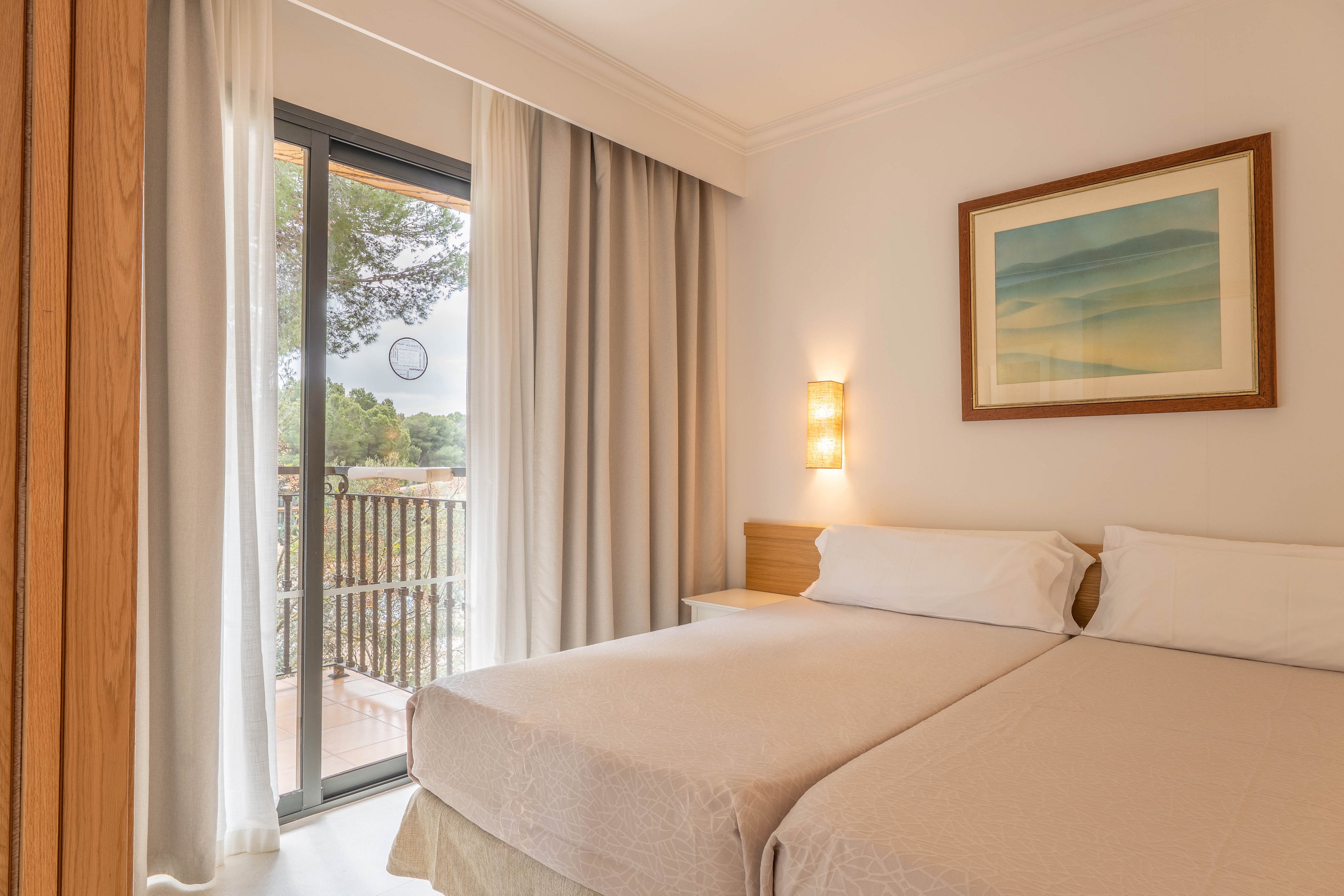 A cozy hotel bedroom features two neatly made single beds with light-colored linens. Soft wall lighting and a framed abstract seascape add warmth to the neutral decor. Large sliding glass doors open to a balcony with a railing and trees visible outside. Floor-to-ceiling curtains frame the doorway, creating an inviting and tranquil atmosphere.