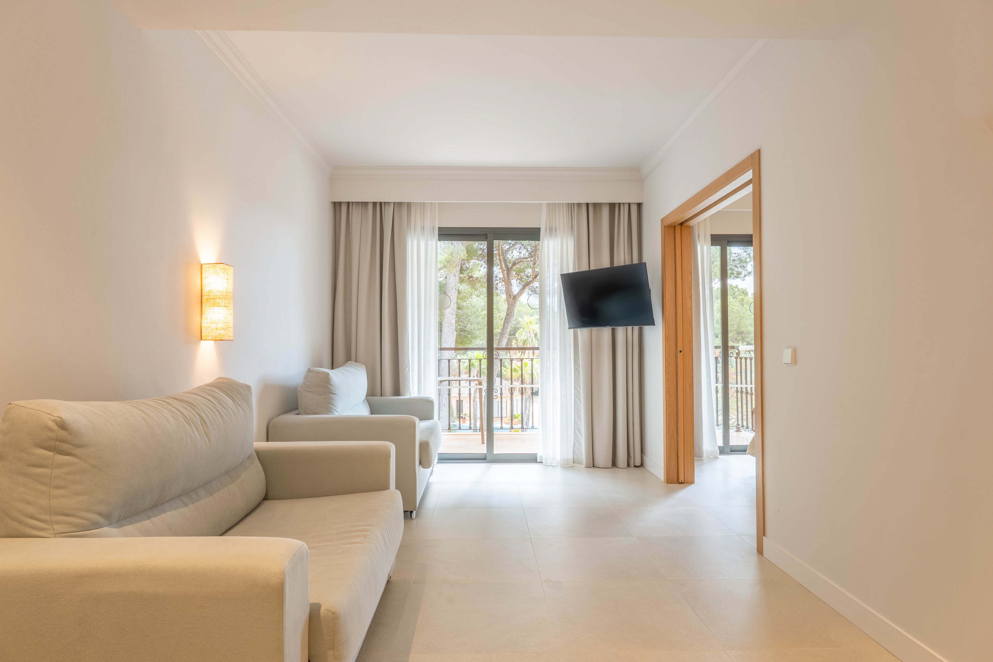 A bright modern living room features two light-colored sofas facing a wall-mounted television. Large sliding glass doors open onto a balcony with a view of trees outside. Neutral walls, floor tiles, and soft curtains create a calm, minimalist atmosphere. A warm wall sconce adds subtle lighting to the cozy seating area.