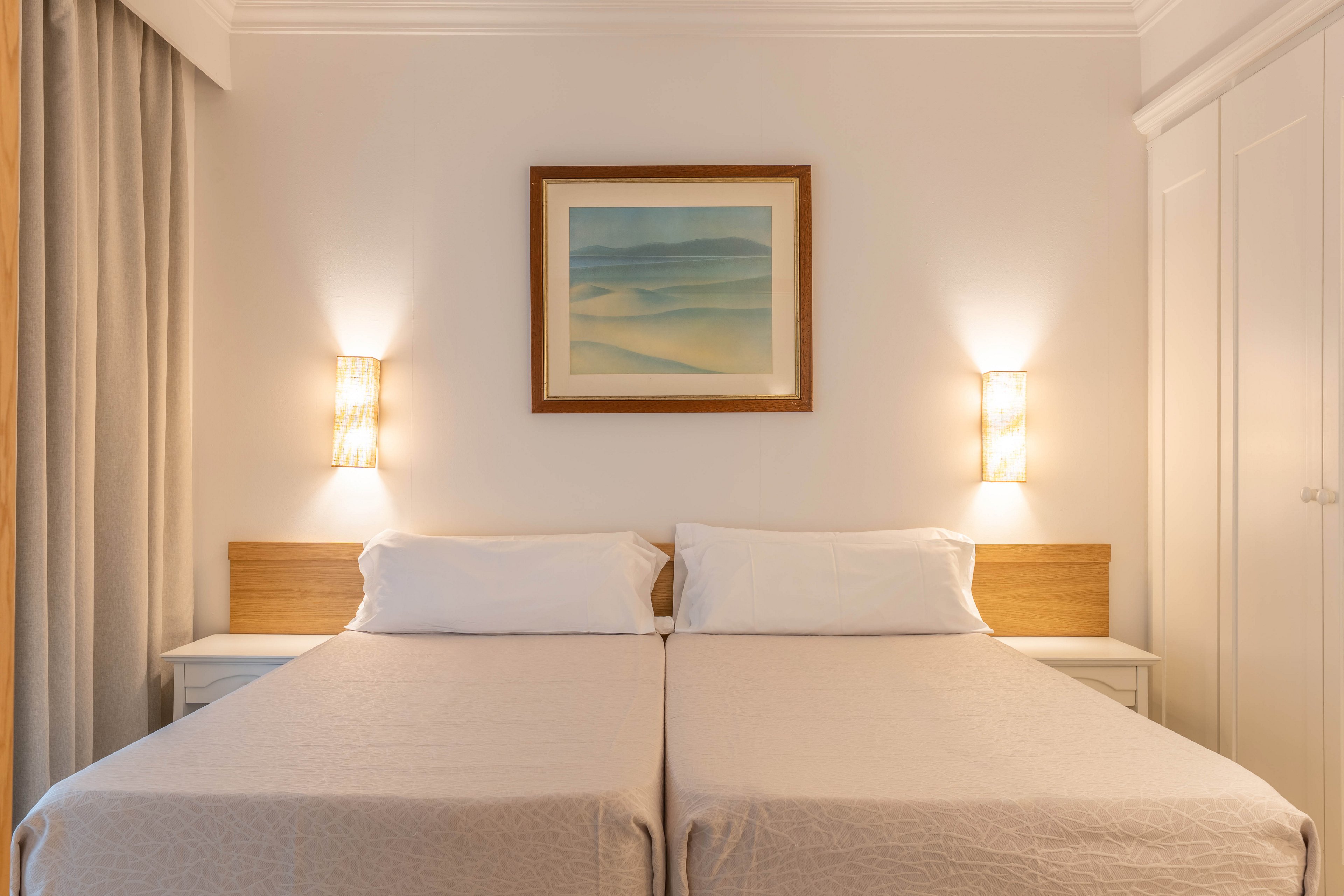 A pair of neatly made twin beds sits side by side against a light-colored wall in a minimalist bedroom. Warm wall sconces on either side of a framed abstract landscape provide soft, ambient lighting. Neutral bedding and curtains create a calm, inviting atmosphere. Built-in wardrobes and bedside tables suggest a tidy, functional hotel or guest room setting.