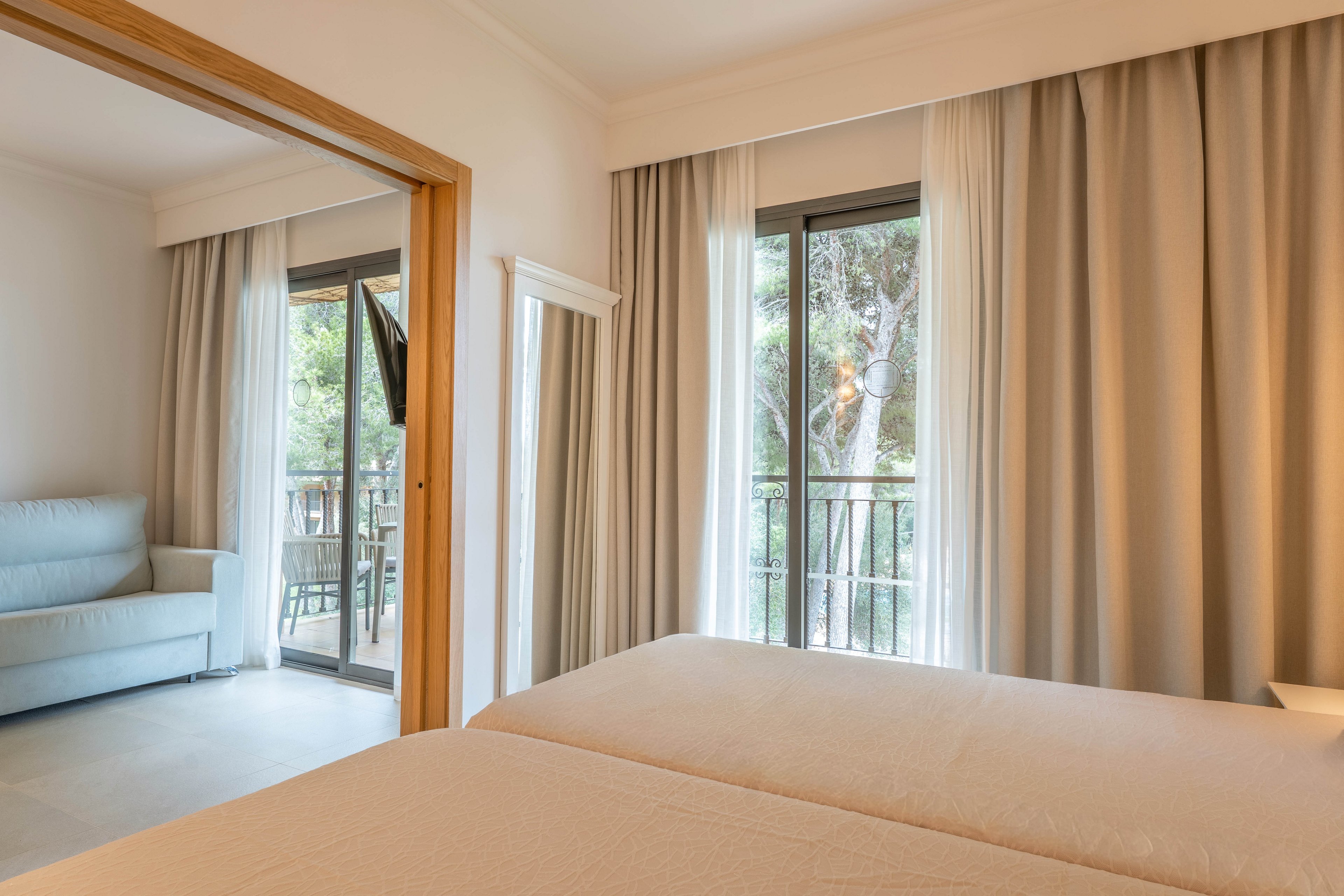 A modern hotel bedroom features twin beds with light-colored linens. Large sliding glass doors open to a balcony overlooking green trees. Neutral curtains and walls create a calm, minimalist atmosphere. A separate sitting area with a light sofa is visible through a wide doorway, enhancing the sense of space and comfort.