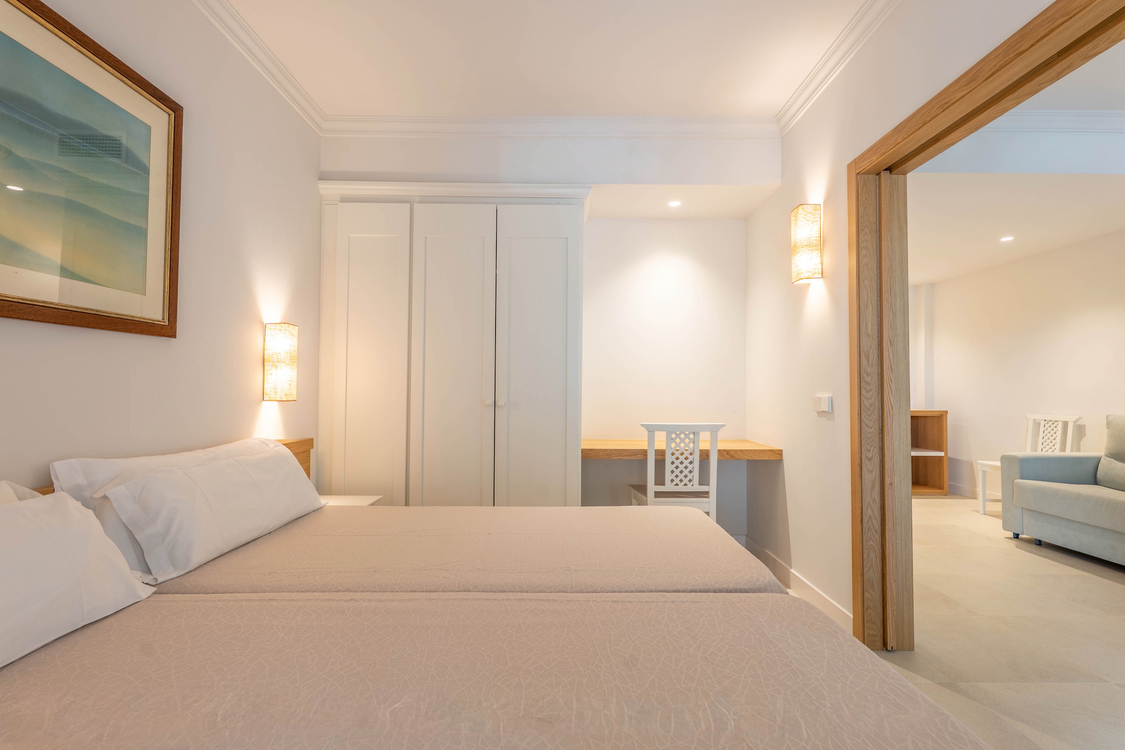 A bright, minimalist bedroom features two neatly made beds with light-colored linens. Built-in white wardrobes and a simple desk with a chair sit against the far wall, illuminated by modern wall sconces. An open wooden-framed doorway leads to an adjoining living area with a sofa and additional seating. Soft neutral tones and clean lines create a calm, contemporary atmosphere suitable for a hotel or vacation rental.
